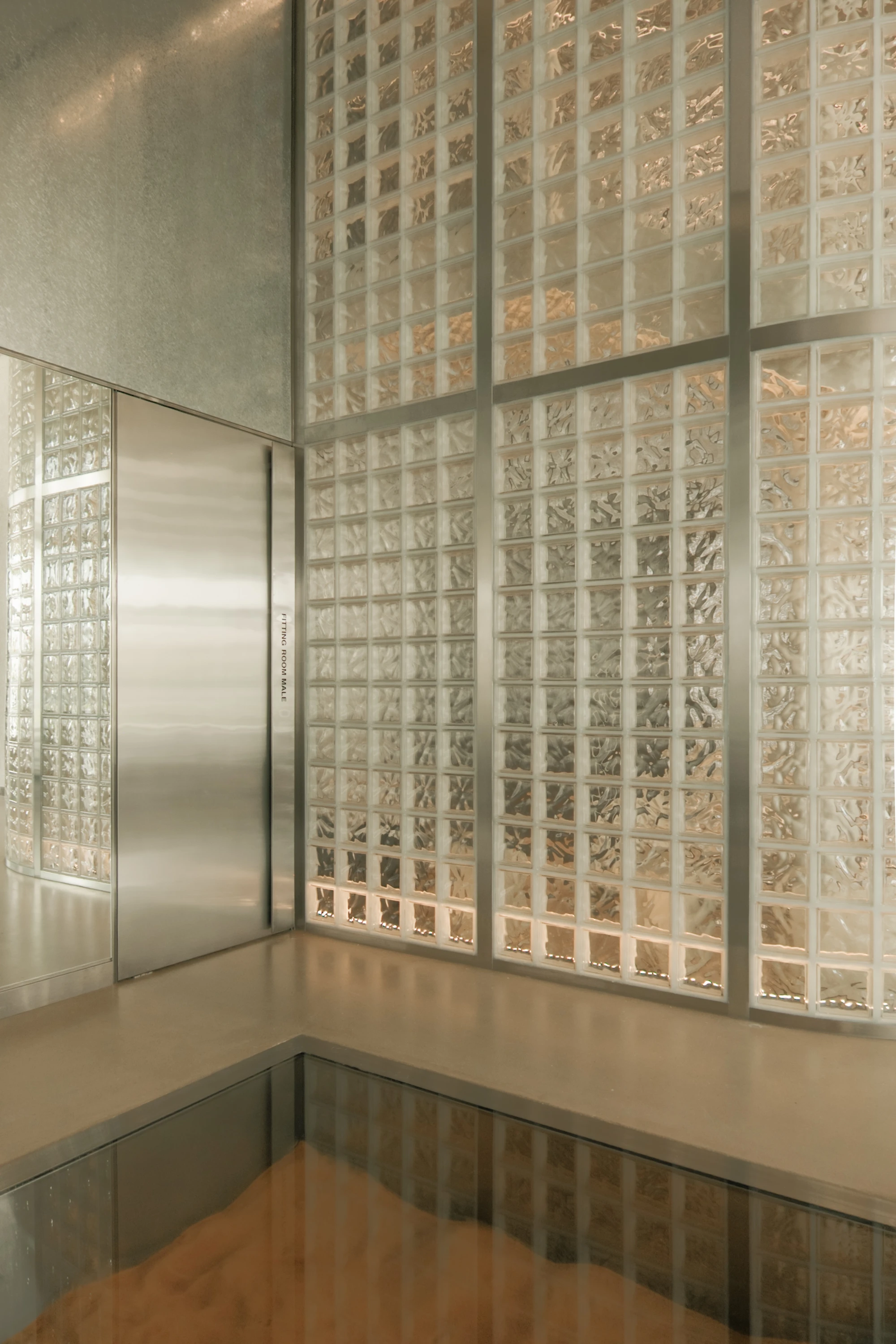 OUNASS STAGE by VAUST, photographed by Nicolas Quiniou — close view of a stainless steel changing-room door set into the glass-block silo wall, with a sand pit sealed under walkable glass visible in the polished concrete floor.