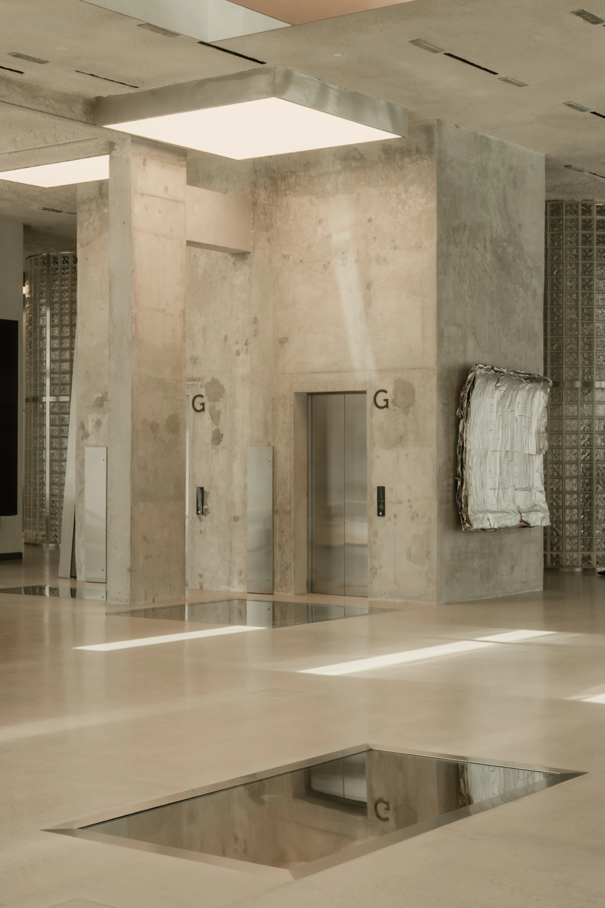 OUNASS STAGE by VAUST, photographed by Nicolas Quiniou — two recessed sand pits sealed under walkable glass set into the polished concrete slab, framed by board-formed concrete columns with stencilled letter G markings and a foil tapestry mounted to the r