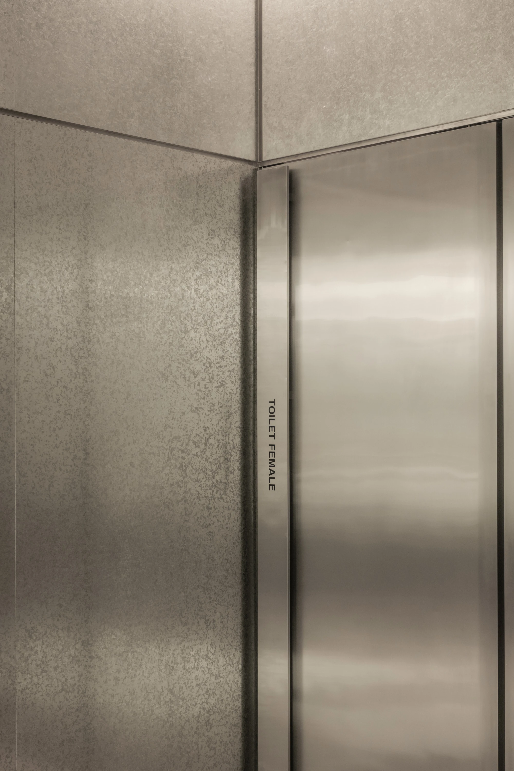 OUNASS STAGE by VAUST, photographed by Nicolas Quiniou — detail of a stainless steel toilet door in the rear wall with a hairline engraved Toilet Female label set vertically into the frame, surrounded by mottled galvanized steel cladding.