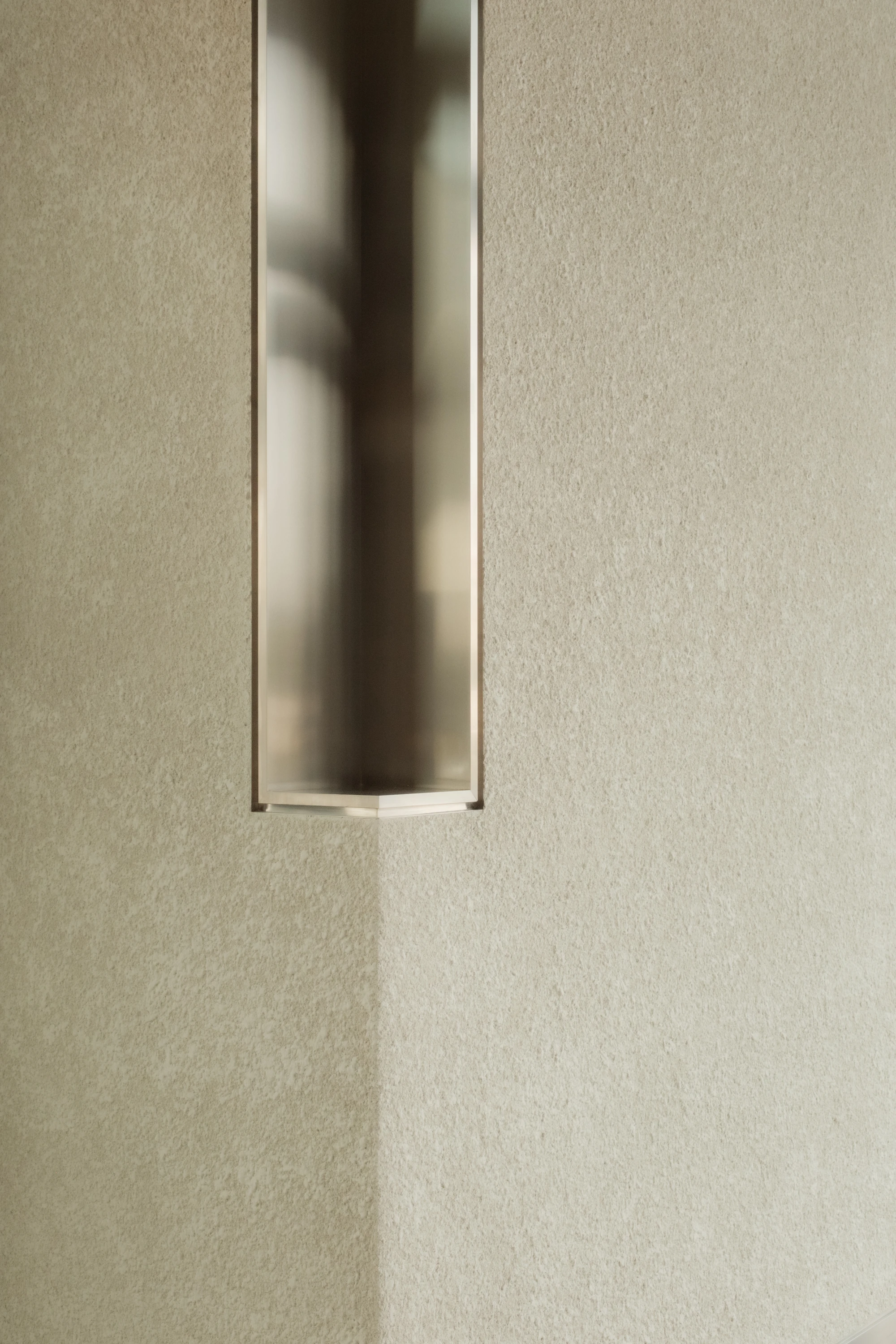 OUNASS STAGE by VAUST, photographed by Nicolas Quiniou — vertical stainless steel slot recess set into a sand-coloured plaster wall, the polished metal catching reflections from above.
