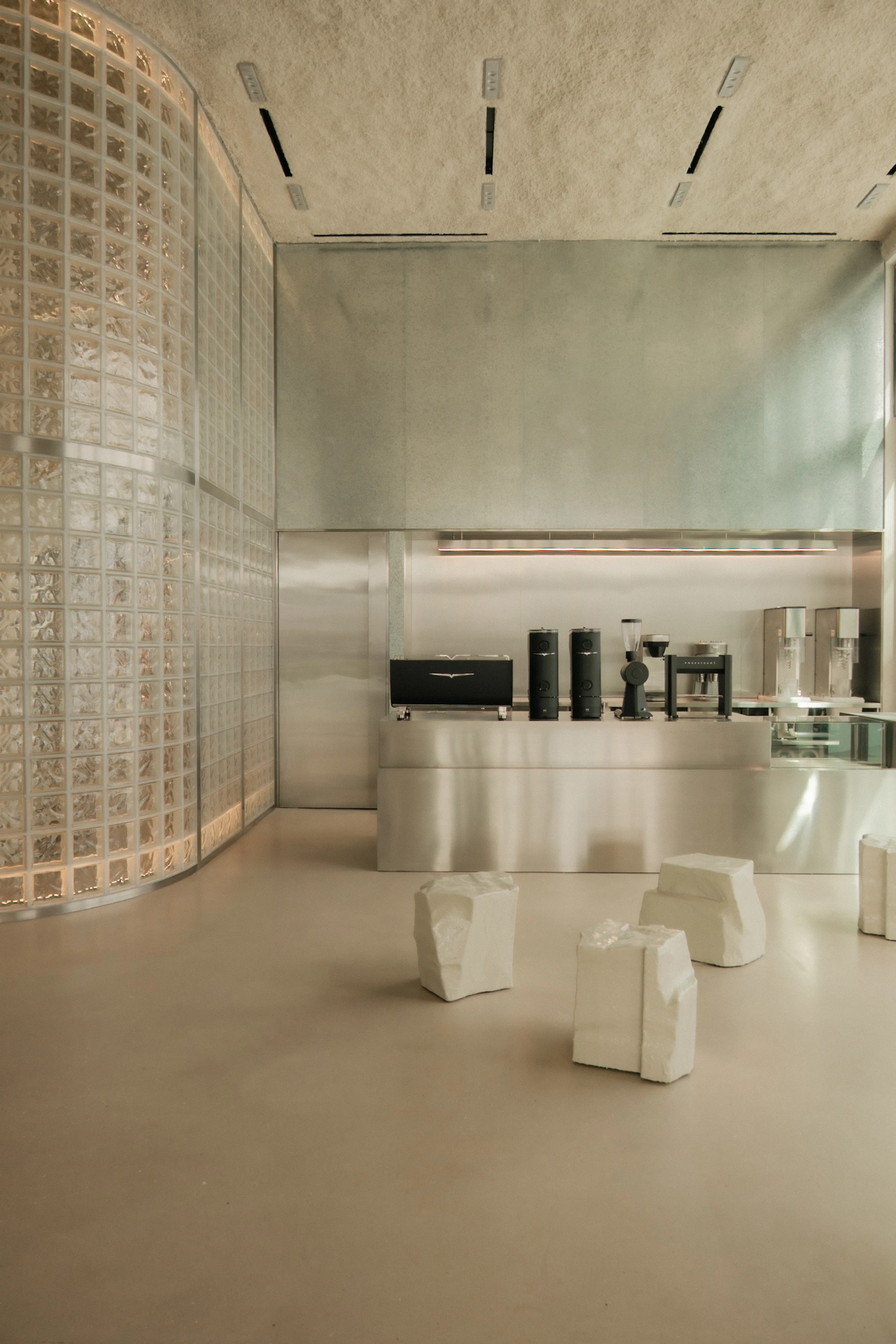 OUNASS STAGE by VAUST, photographed by Nicolas Quiniou — the stainless steel coffee bar with grouped objects on its counter set beside the glass-block silo, with white faceted plinths arranged on the polished concrete floor.