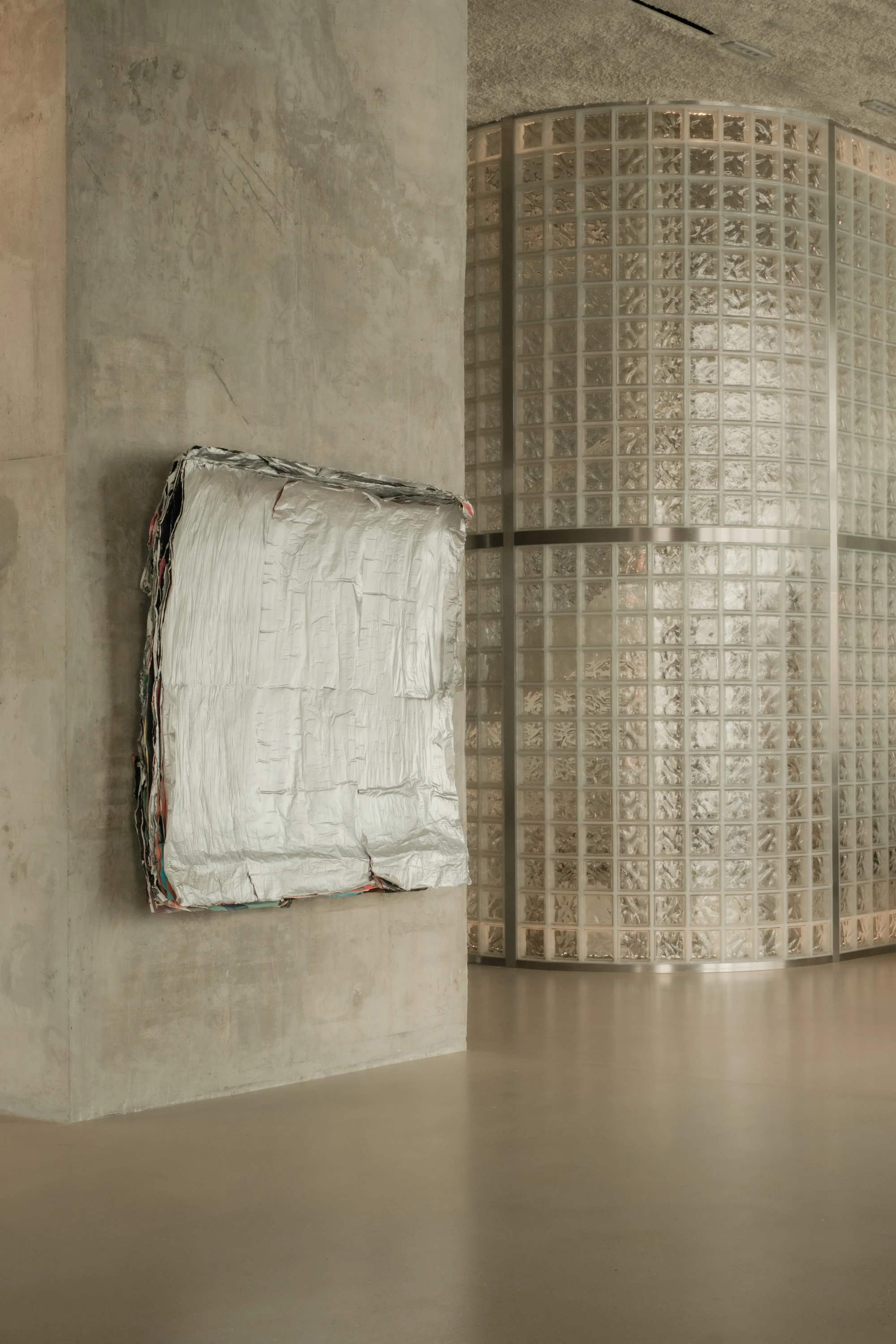 OUNASS STAGE by VAUST, photographed by Nicolas Quiniou — Joern von Scheipers' crumpled-foil tapestry hanging flat against a board-formed concrete column, with a curved glass-block silo backlit beside it.