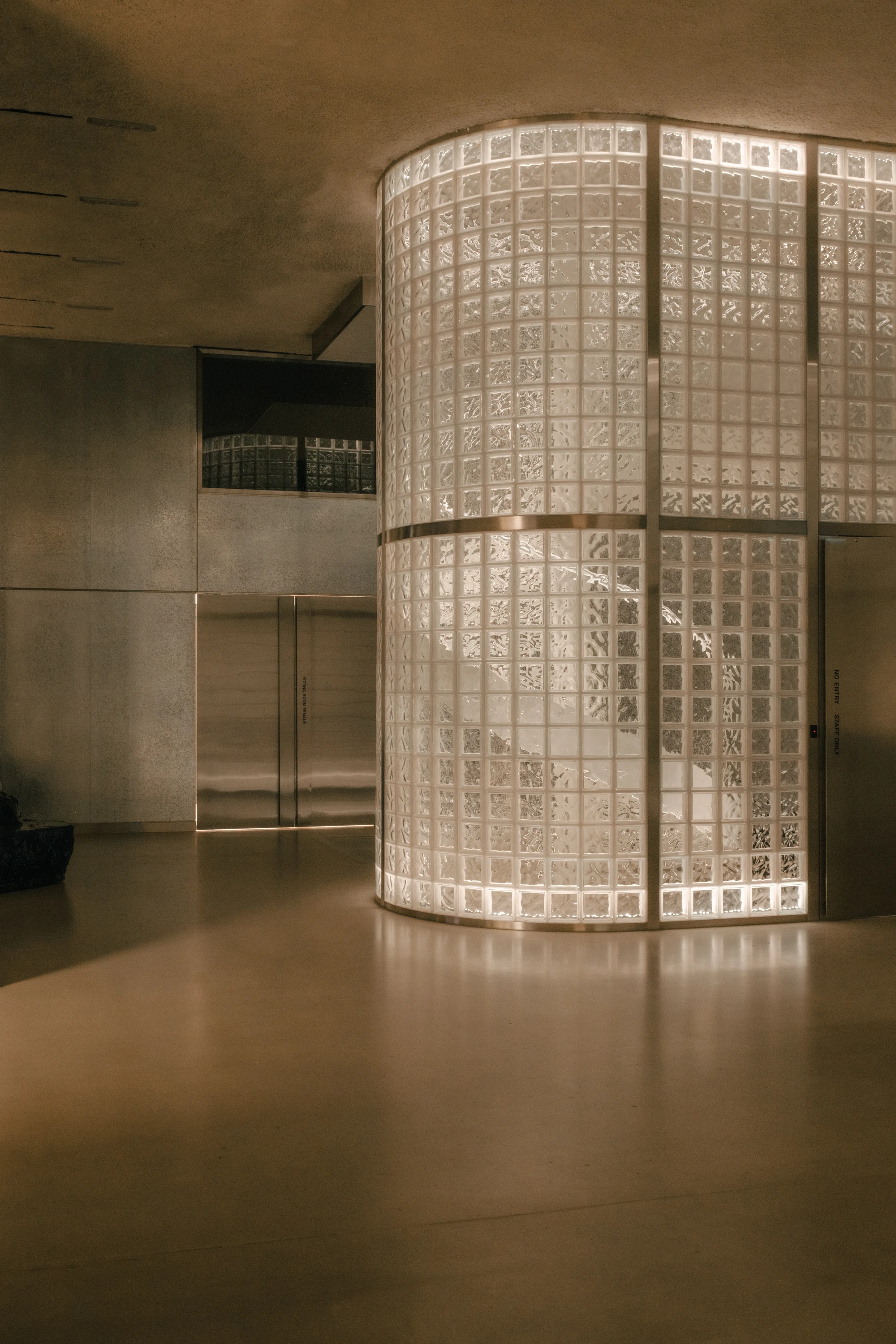 OUNASS STAGE by VAUST, photographed by Nicolas Quiniou — a backlit cylindrical glass-block silo glowing amber at dusk, set against the dark galvanized steel rear wall and the stainless steel service doors of the inhabited infrastructure spine.