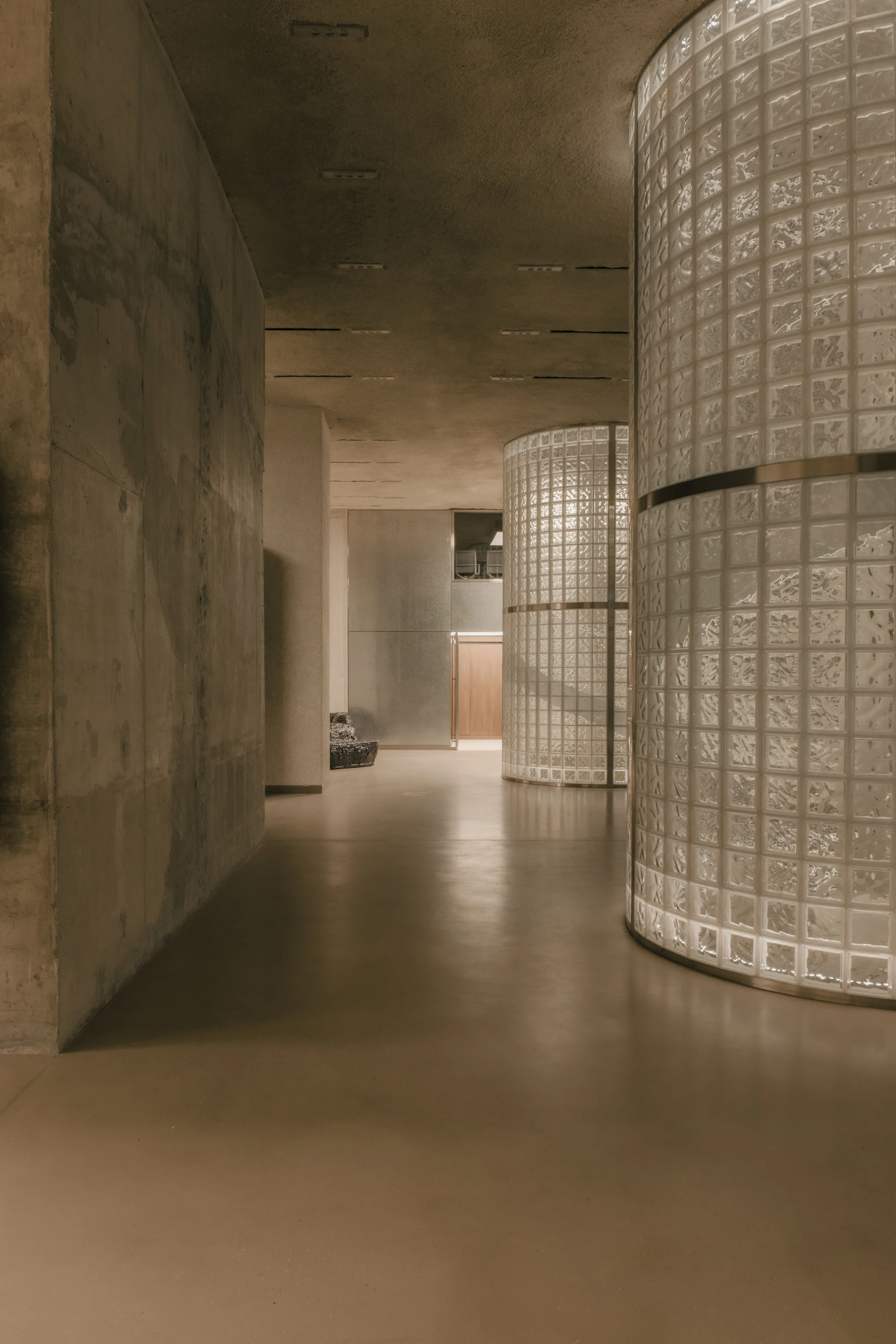 OUNASS STAGE by VAUST, photographed by Nicolas Quiniou — a circulation corridor where board-formed concrete meets a row of curved glass-block silos lit from within, the polished concrete floor reflecting their warm glow.
