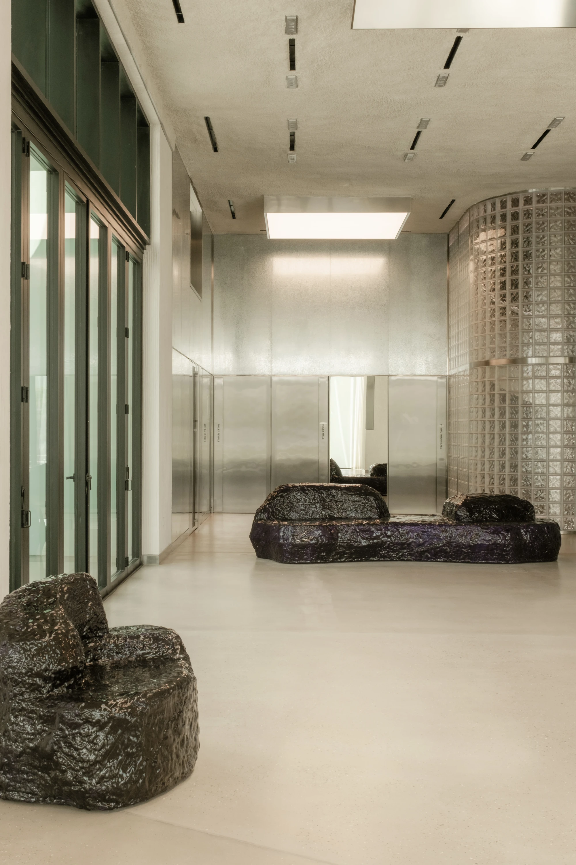 OUNASS STAGE by VAUST, photographed by Nicolas Quiniou — the galvanized steel rear wall reflecting overhead skylights, with two boulder-form Balzer Balzer sofas set on polished concrete in front of a glass-block silo.