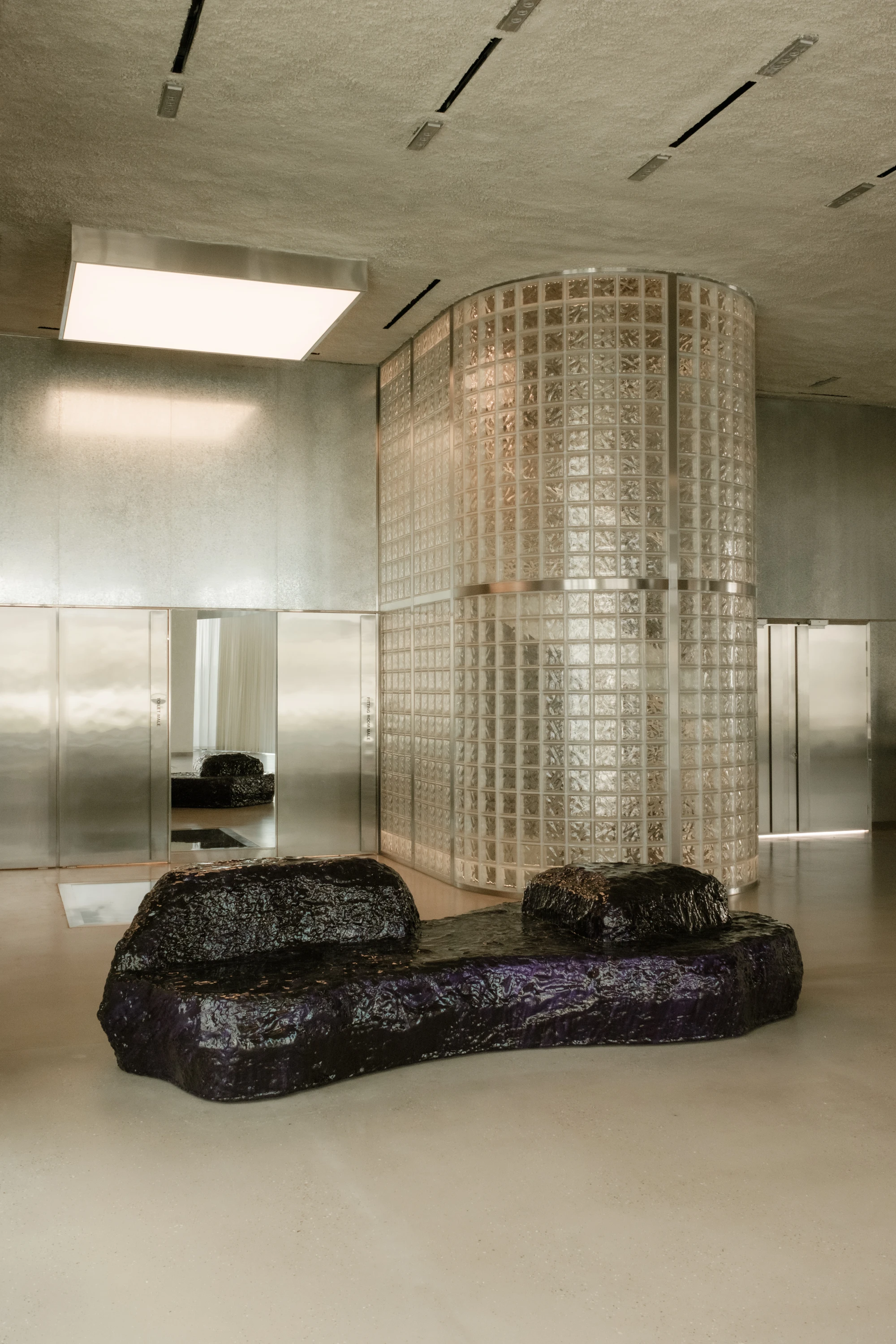 OUNASS STAGE by VAUST, photographed by Nicolas Quiniou — frontal view of a cylindrical glass-block changing-room silo flanked by polished galvanized steel rear walls, with a Balzer Balzer boulder sofa set on the polished concrete floor.
