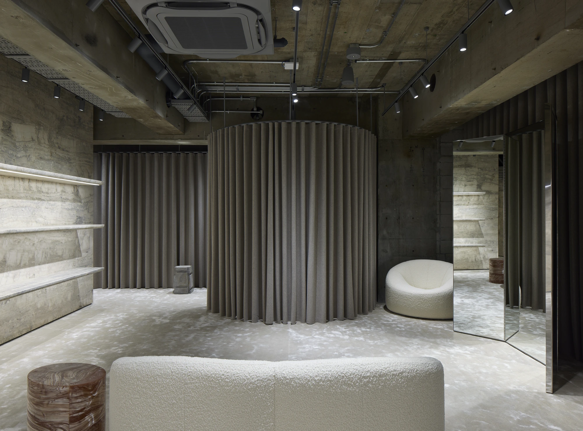 Jil Sander Flagship Store Ginza by Casper Mueller Kneer Architects, photographed by Paul Riddle — upper-floor fitting area with felt curtain drum, cream boucle armchair, mirrored panel, and marbled timber stool on a textured aggregate floor.