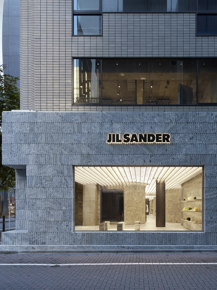 Jil Sander Flagship Store Ginza by Casper Mueller Kneer Architects, photographed by Paul Riddle — the Ginza street facade, chiselled grey-green granite under the Jil Sander signage, gallery window glowing onto the pavement at dusk.