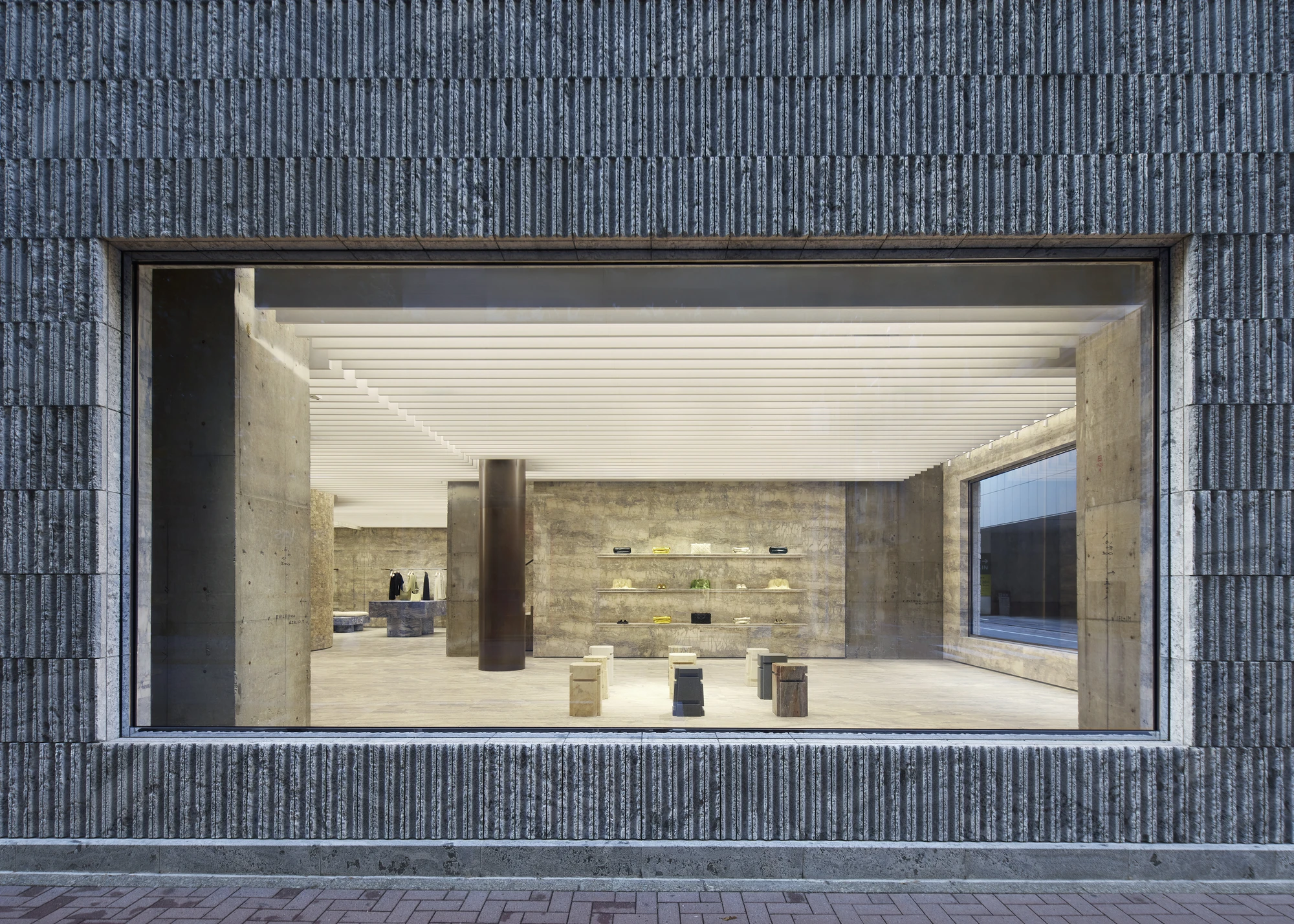 Jil Sander Flagship Store Ginza by Casper Mueller Kneer Architects, photographed by Paul Riddle — frontal street view through the gallery-scale window, board-formed concrete walls and travertine shelving framing the interior.