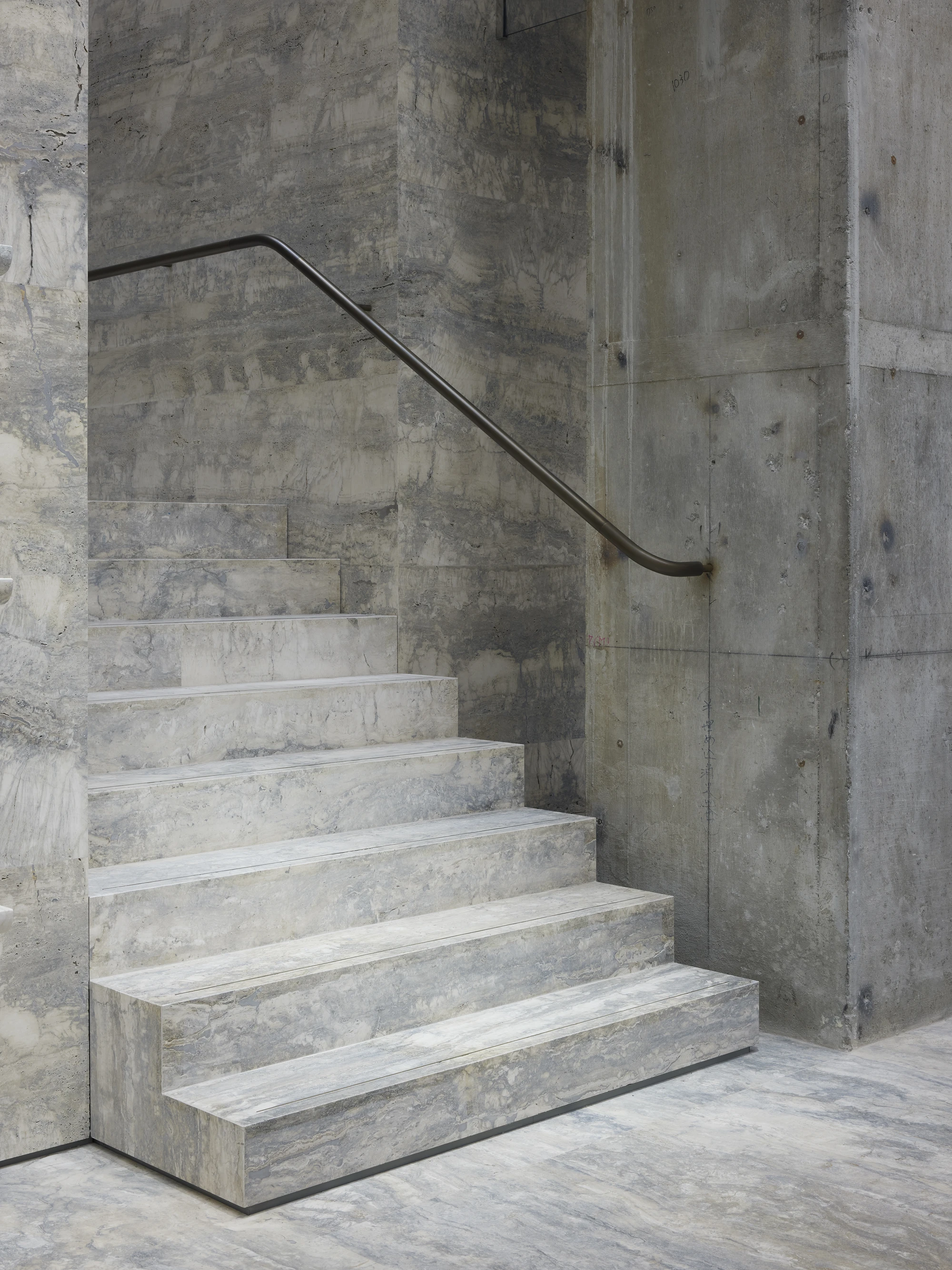 Jil Sander Flagship Store Ginza by Casper Mueller Kneer Architects, photographed by Paul Riddle — silver-blue travertine staircase with a patinated brass handrail tracing the board-formed concrete wall.