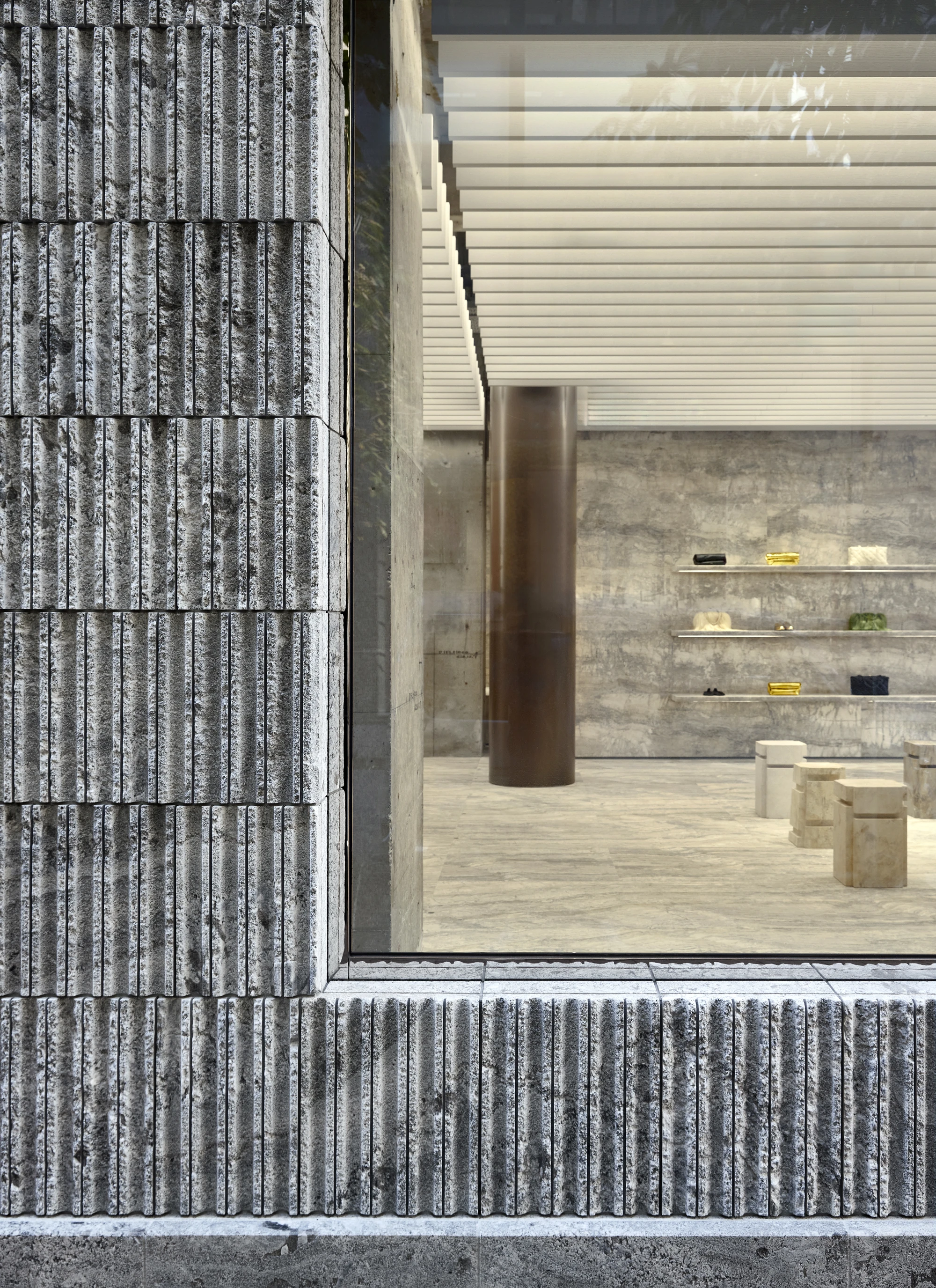 Jil Sander Flagship Store Ginza by Casper Mueller Kneer Architects, photographed by Paul Riddle — granite plinth wall outside a gallery-scale window, with the interior travertine floor and brass column visible beyond.