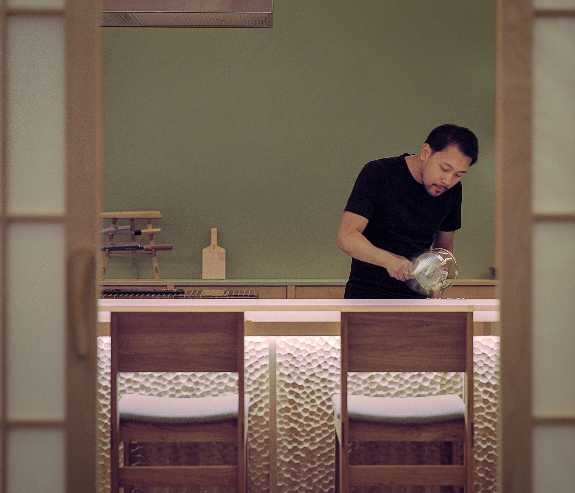 Hos Bobby by NÆRSTUDIO, photographed by Hugo Lütcherath — Chef Bobby Buaprakone at work behind the counter, framed by sliding shoji-style screens and a sage-green wall.