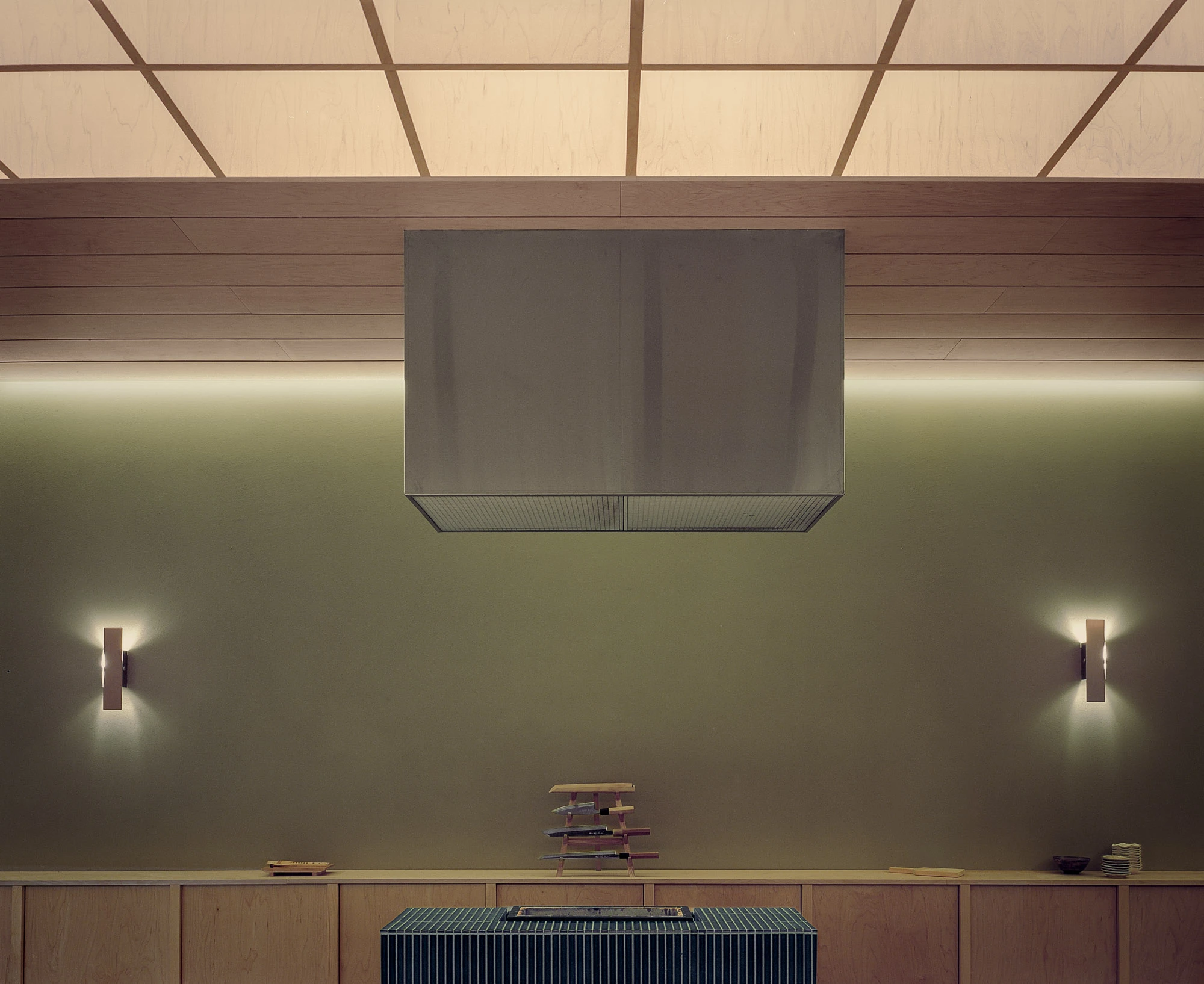 Hos Bobby by NÆRSTUDIO, photographed by Hugo Lütcherath — A stainless extraction hood centers the kitchen wall, flanked by paired sconces beneath the backlit timber ceiling.