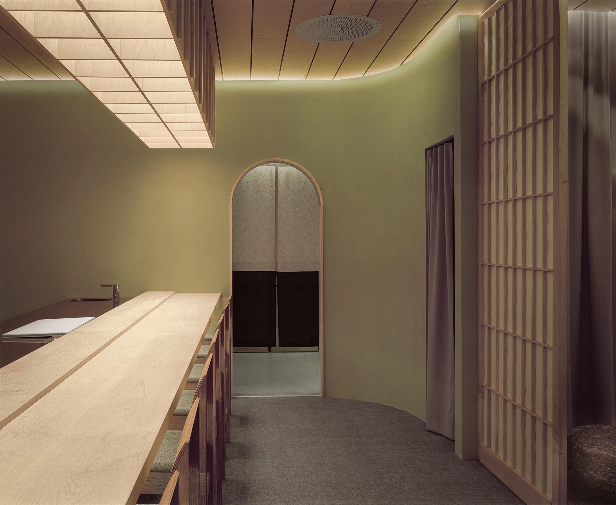 Hos Bobby by NÆRSTUDIO, photographed by Hugo Lütcherath — The omakase counter runs toward an arched doorway hung with a split noren curtain, sliding screens folding the room's edge.