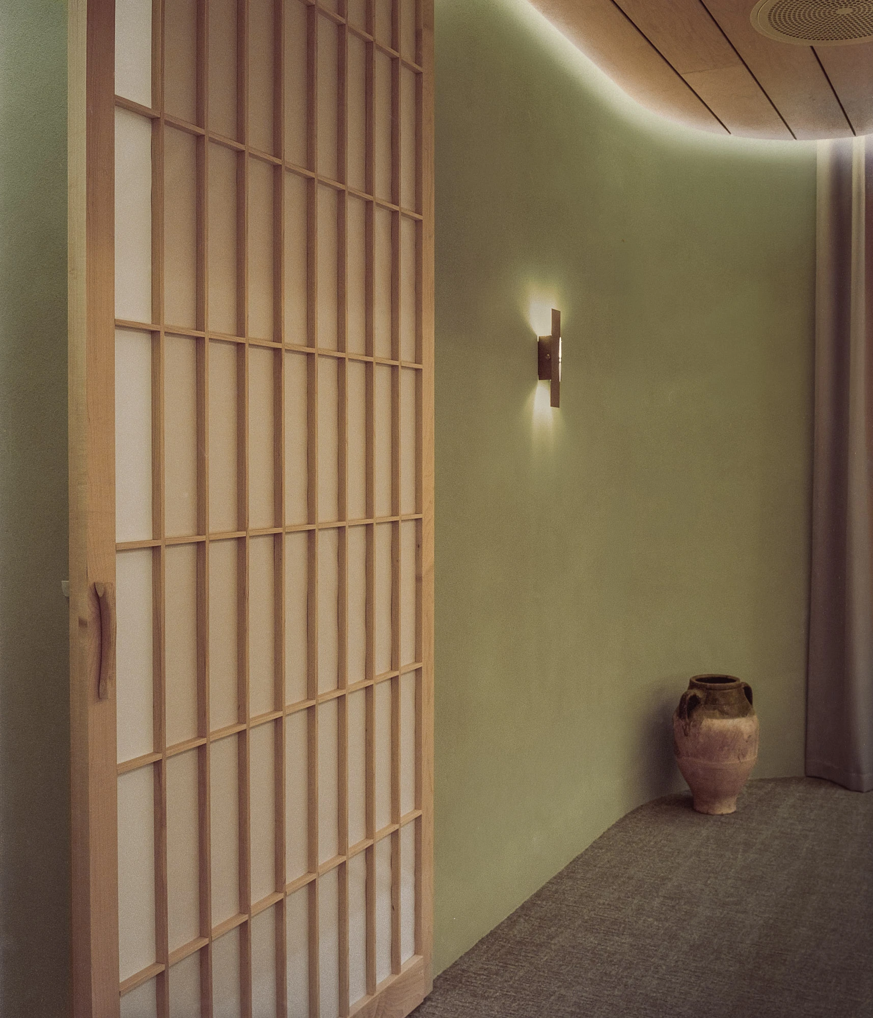 Hos Bobby by NÆRSTUDIO, photographed by Hugo Lütcherath — A sliding screen of slender timber lattice and translucent infill meets the curved clay-plastered wall, a terracotta urn on the carpet.