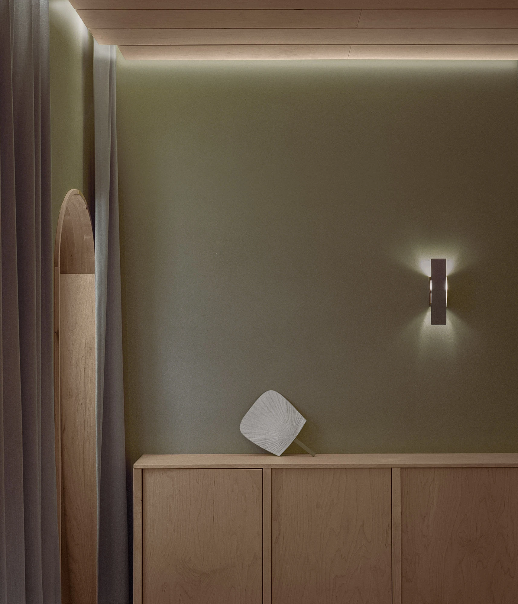 Hos Bobby by NÆRSTUDIO, photographed by Hugo Lütcherath — A paper fan rests on a low timber sideboard beneath a steel wall sconce, the clay plaster washed in cove light.