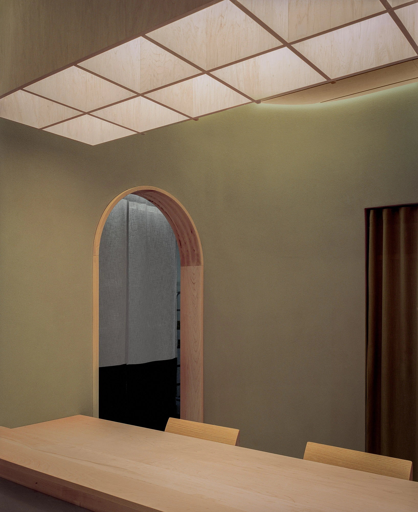 Hos Bobby by NÆRSTUDIO, photographed by Hugo Lütcherath — A coffered grid of backlit veneer panels hovers above the dining table, beside an arched, timber-lined doorway.