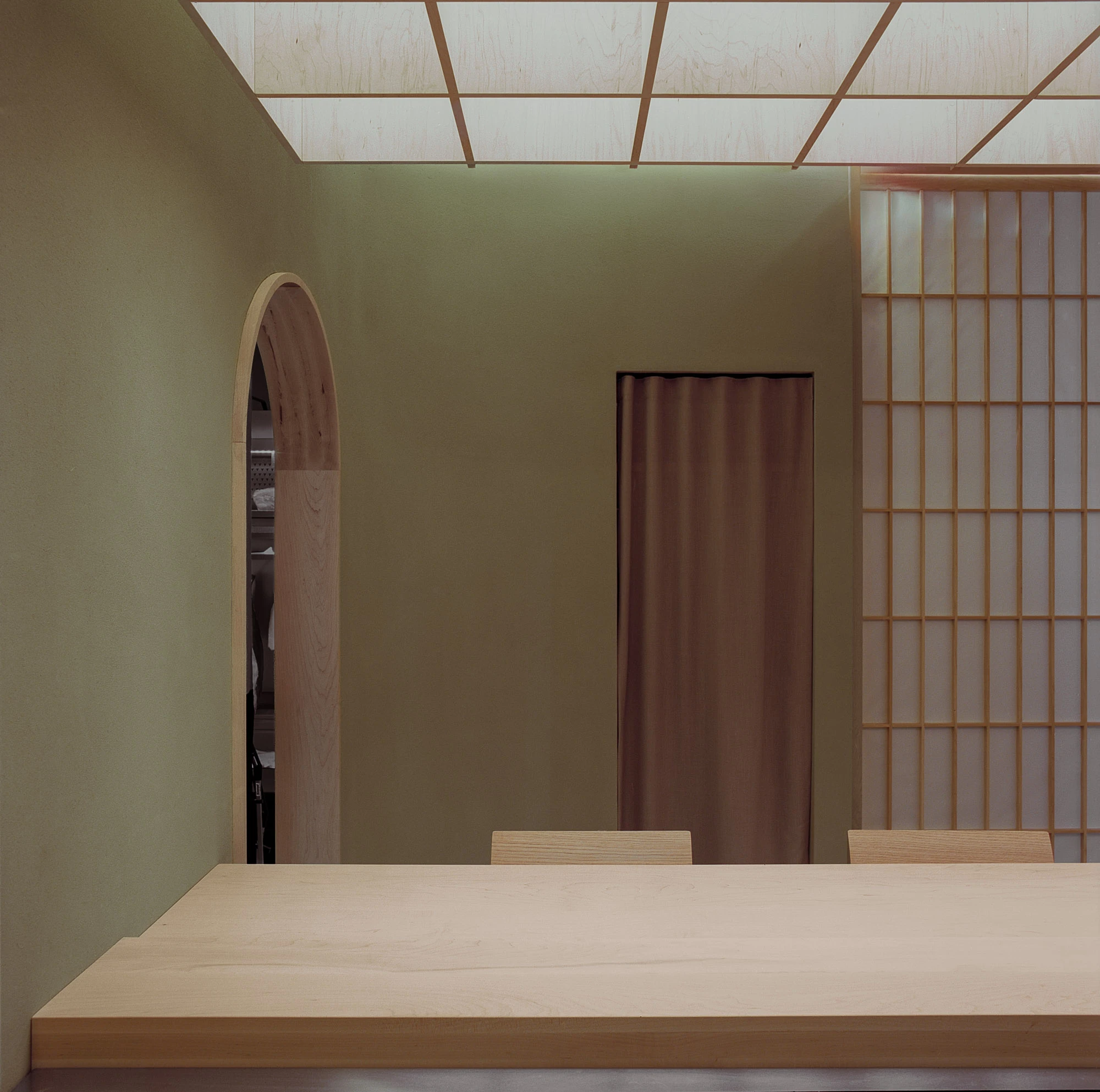 Hos Bobby by NÆRSTUDIO, photographed by Hugo Lütcherath — The dining room's pale timber table under the backlit ceiling grid, an arched opening cut into green clay plaster.