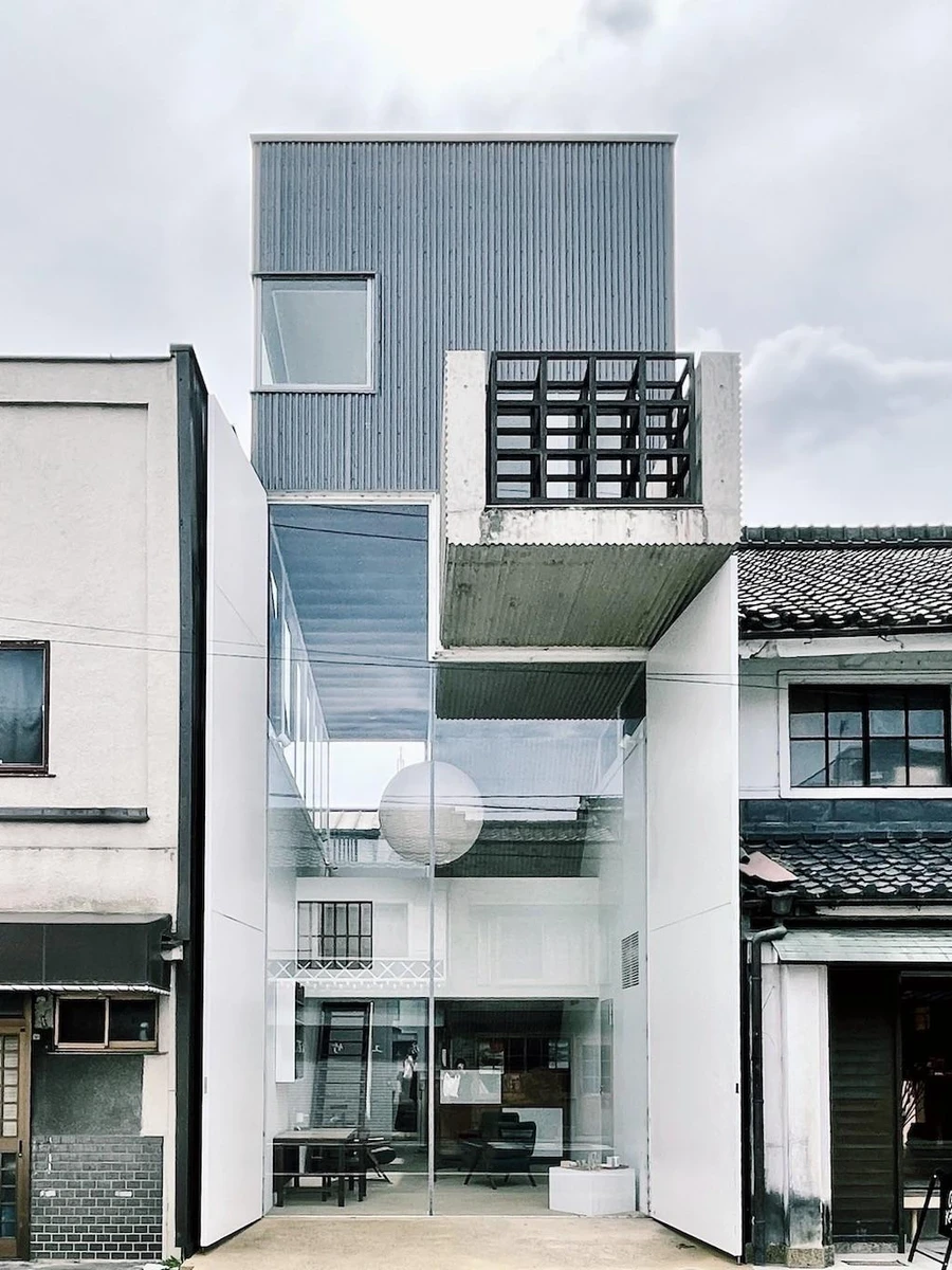 AA Okayama by Jonathan Monk + Go Hasegawa, a frontal street view of the three-volume hotel, with a glass facade flanked by a tiled cafe and a stuccoed neighbour.