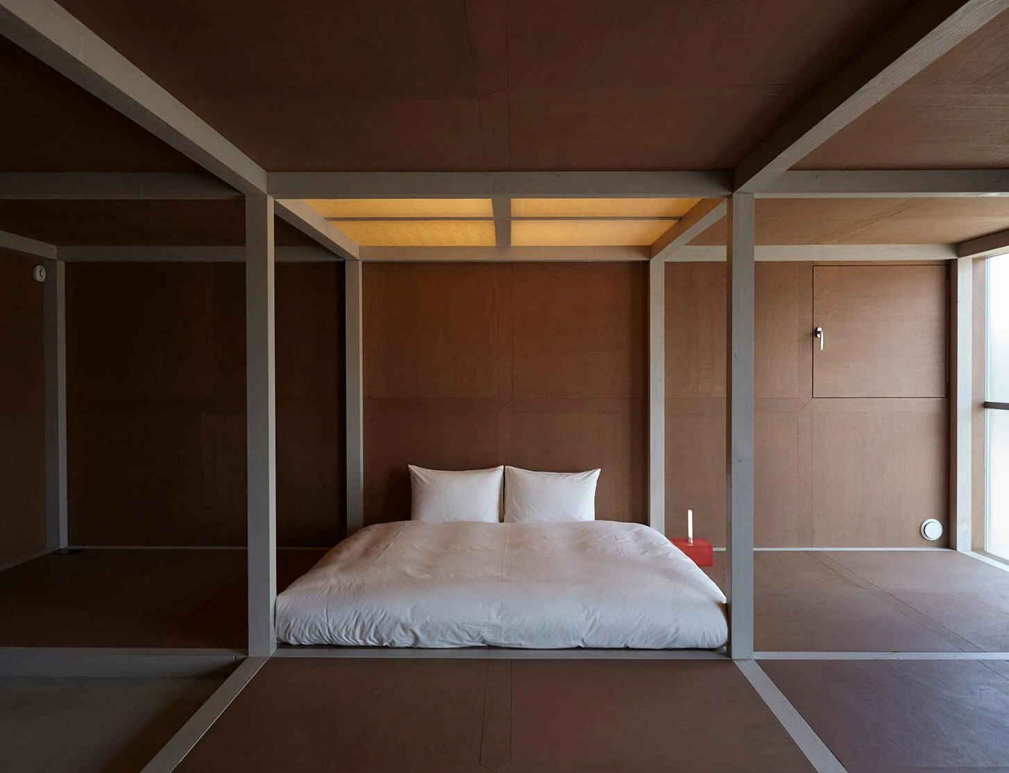 AA Okayama by Jonathan Monk + Go Hasegawa, a futon laid inside a plywood-lined sleeping alcove, with a red lacquered side box holding a single candle.