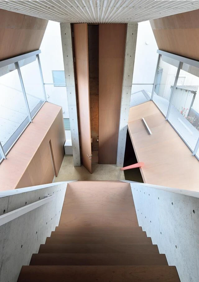 AA Okayama by Jonathan Monk + Go Hasegawa, an overhead view down a plywood and concrete stair toward an open timber door, with adjoining buildings visible through floor-to-ceiling glass.