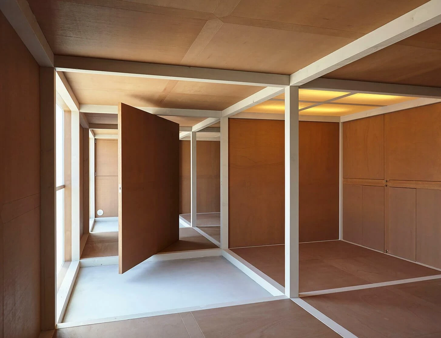 AA Okayama by Jonathan Monk + Go Hasegawa, a plywood-lined interior framed by slender white-painted timbers, with a pivoting door and an internal window glowing yellow above.
