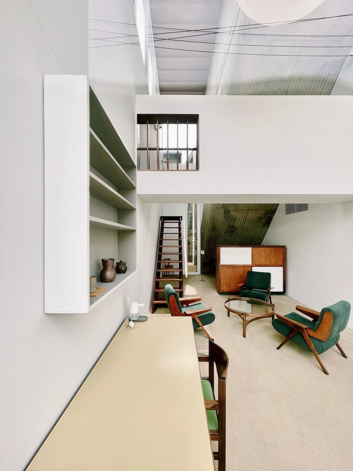 AA Okayama by Jonathan Monk + Go Hasegawa, the double-height ground-floor lounge with a fixed wall shelf of ceramics, green velvet armchairs and a steep wooden ladder leading upward.