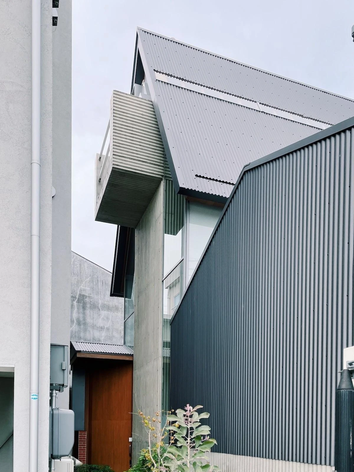 AA Okayama by Jonathan Monk + Go Hasegawa, a side view of the central volume, where corrugated metal folds from wall into pitched roof over a concrete cantilevered balcony.