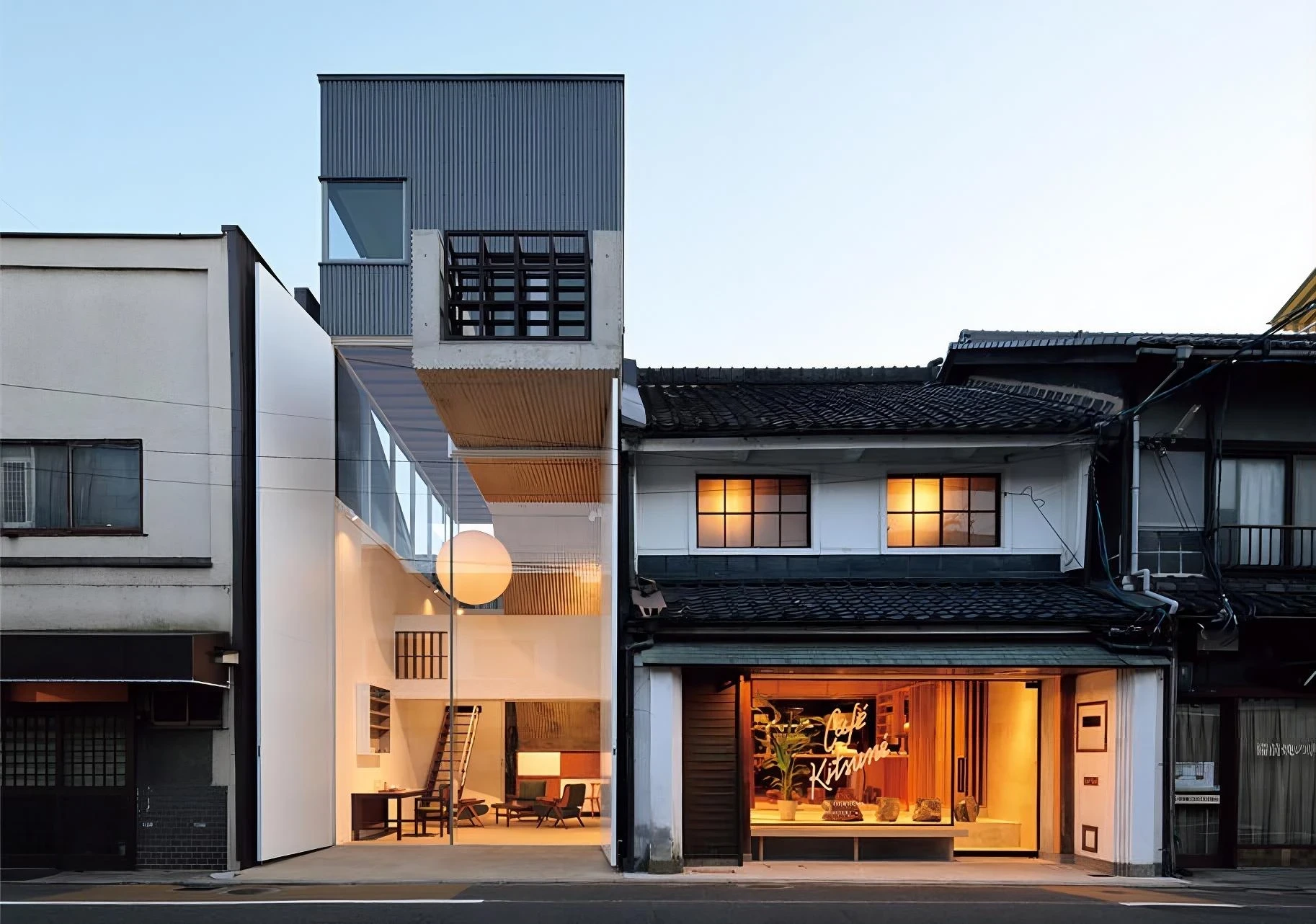 AA Okayama by Jonathan Monk + Go Hasegawa, the three-building hotel at dusk with a glass-fronted ground floor opening onto the street between a corrugated tower and a tiled merchant house.