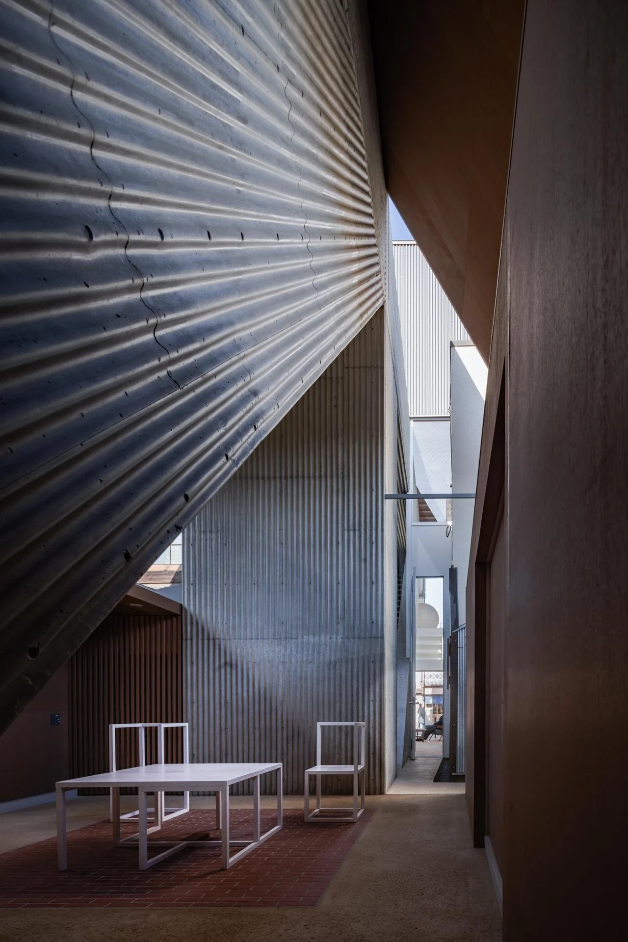 AA Okayama by Jonathan Monk + Go Hasegawa, a brick-paved courtyard inside the hotel where a draped corrugated metal ceiling sags between buildings.