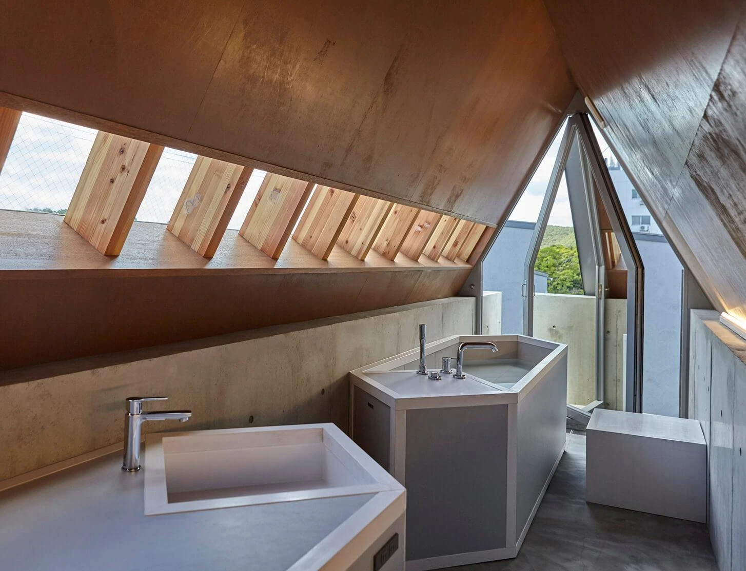 AA Okayama by Jonathan Monk + Go Hasegawa, the top-floor bathroom under the gabled corrugated roof, with cedar louvers, a stone bath and a triangular window cut to follow the gable.