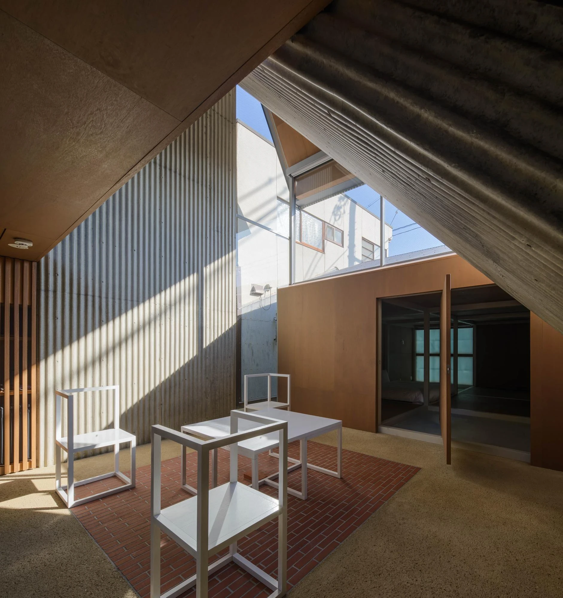 AA Okayama by Jonathan Monk + Go Hasegawa, the daylit courtyard between the three buildings, with white minimal chairs on red brick paving under a corrugated metal roof.