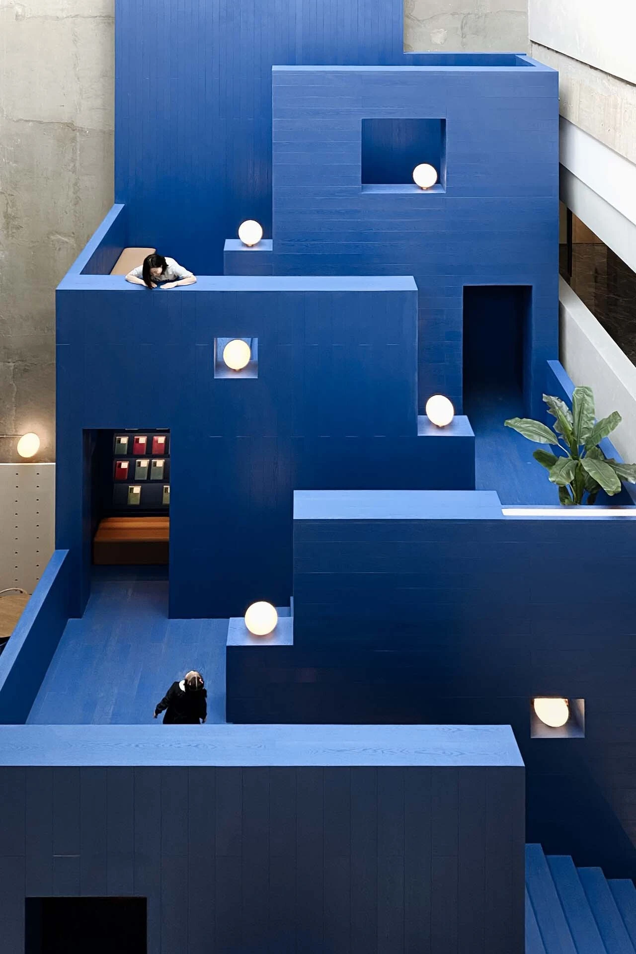 LVWA Bookstore by Studio YUDA + Studio NOR, photographed by CreatAR Images — seen from above, the mountain's stepped blue-oak terraces nest into one another, a reader leaning on a railing on the upper level as the peak retreats in five tiers toward a plan