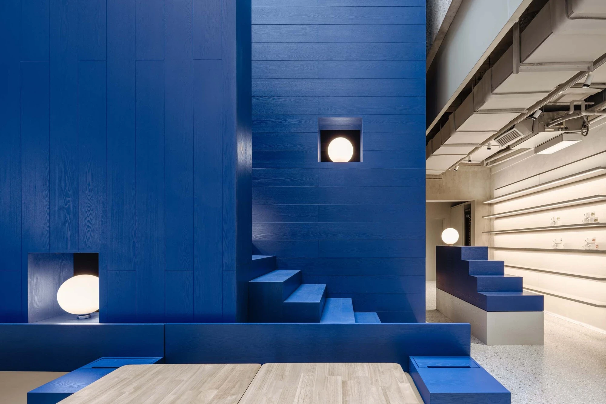 LVWA Bookstore by Studio YUDA + Studio NOR, photographed by CreatAR Images — inside the mountain, a blue-oak stair runs between two walls in cobalt-stained vertical planks, lit by a single recessed globe sconce set into a square wall-niche.