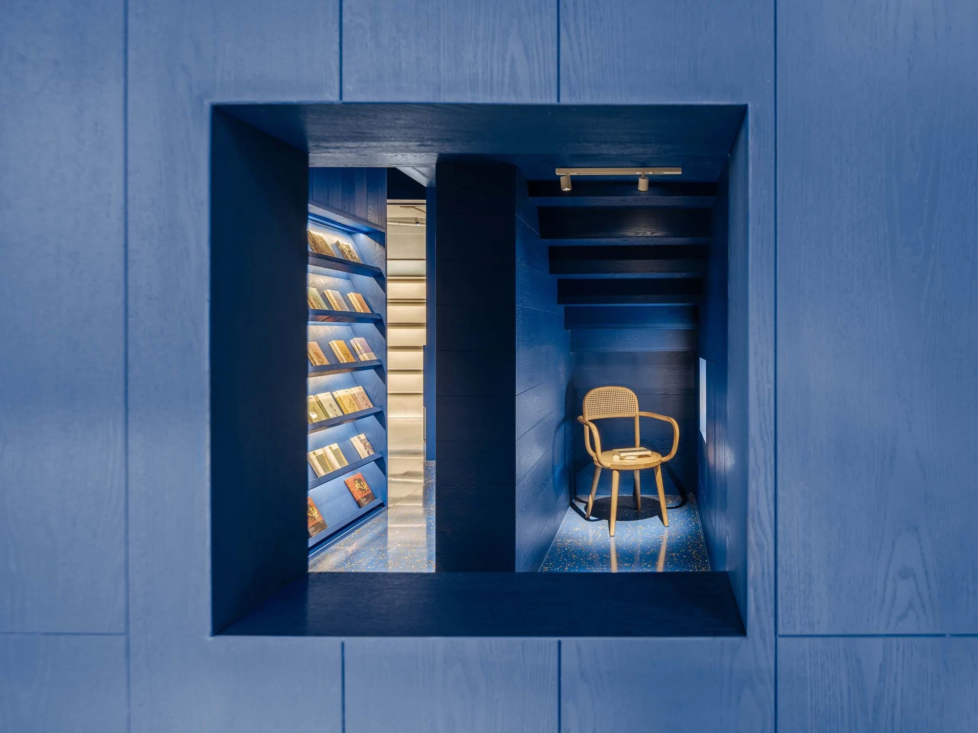 LVWA Bookstore by Studio YUDA + Studio NOR, photographed by CreatAR Images — a square window cut in the mountain frames a reading cell: a single cane-back ash chair on a blue-flecked terrazzo floor, lit by a brass-mounted track light over face-out book di