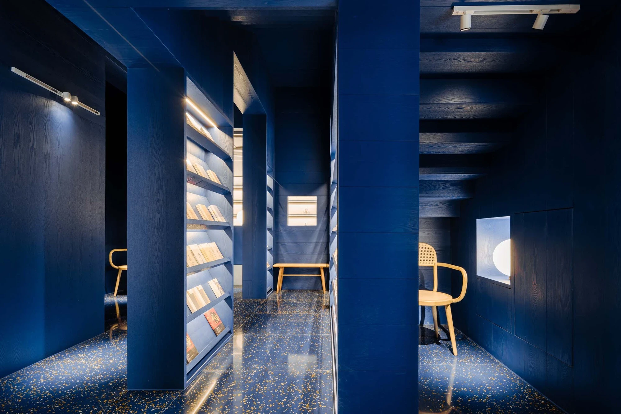 LVWA Bookstore by Studio YUDA + Studio NOR, photographed by CreatAR Images — a passage between blue-oak book mountains: shelves of paperbacks open along the right wall, a perforated metal door catches daylight at the end of the corridor, and the blue-flec