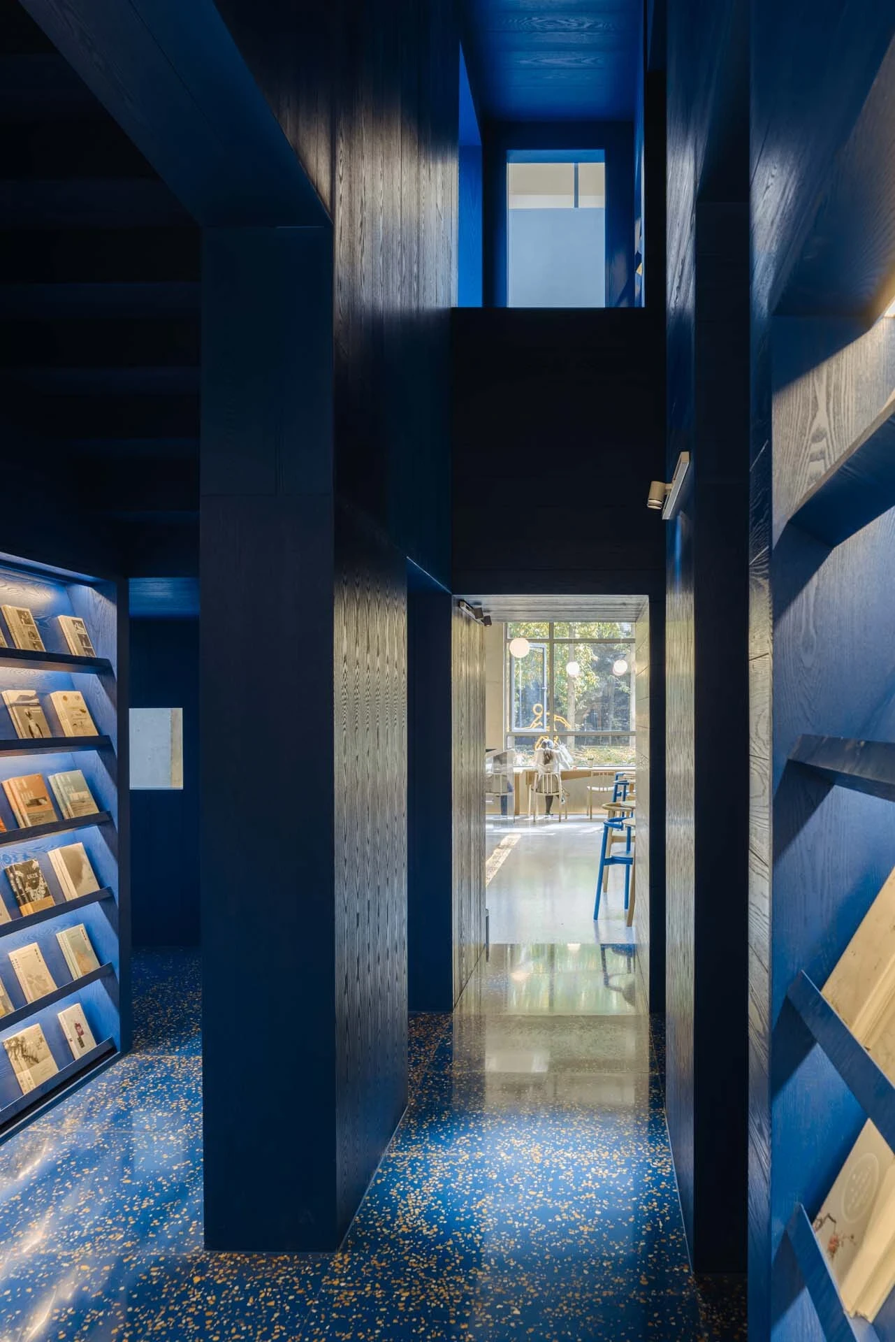 LVWA Bookstore by Studio YUDA + Studio NOR, photographed by CreatAR Images — stepped blue-oak shelving units shape a small reading room, lit by globe sconces and a low pendant; a cane-back chair sits at the rear under a framed indigo print.