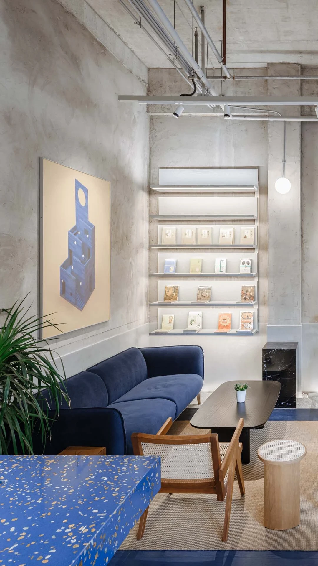 LVWA Bookstore by Studio YUDA + Studio NOR, photographed by CreatAR Images — a quiet lounge alcove: a deep cobalt velvet sofa sits under a framed indigo painting of the mountain, with thin shelves of books climbing a board-formed concrete wall next to a b