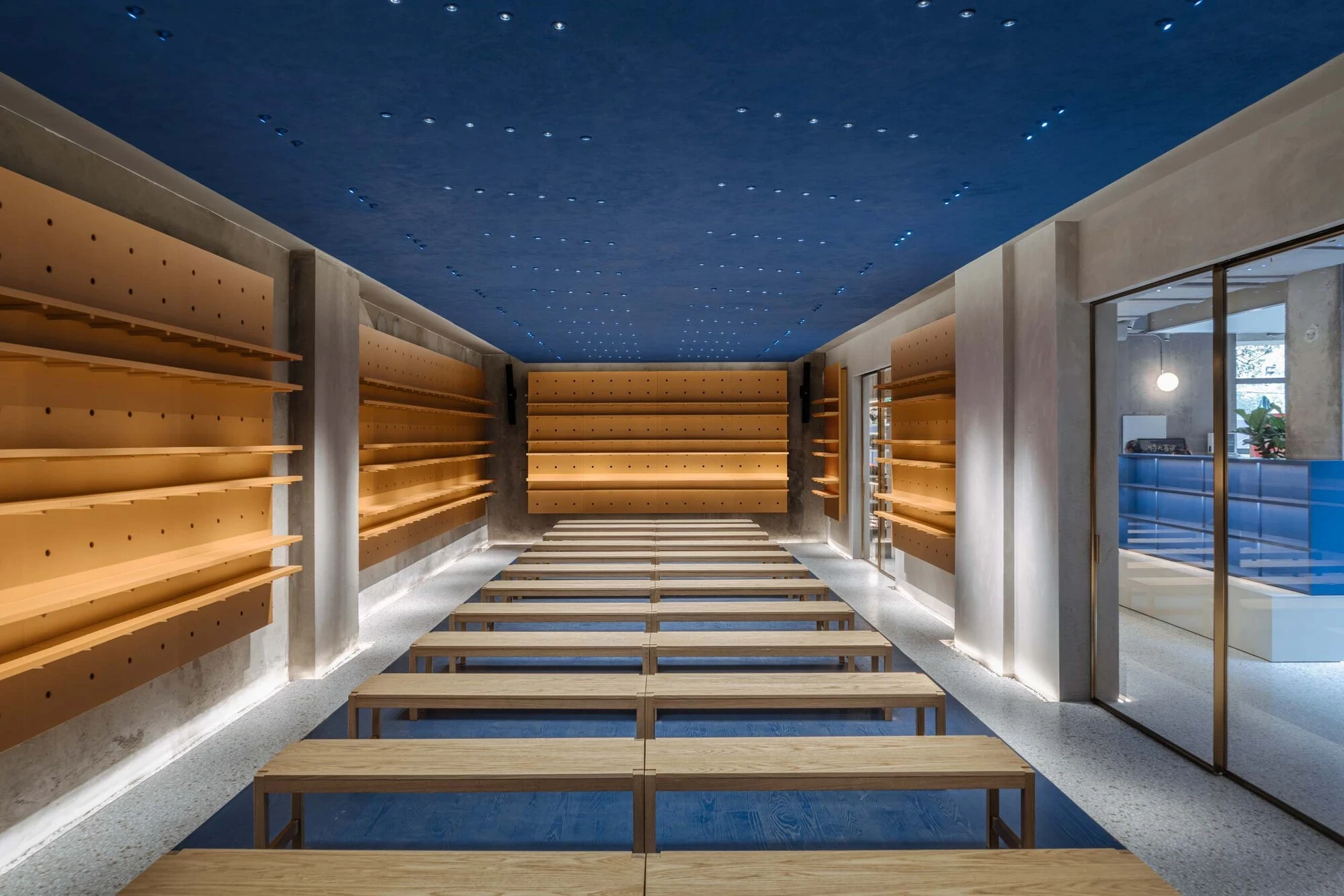 LVWA Bookstore by Studio YUDA + Studio NOR, photographed by CreatAR Images — the lecture hall: rows of pale ash benches sit on a cobalt floor under a blue-felt ceiling speckled with pinpoint downlights, perforated-board shelves climbing both side walls an