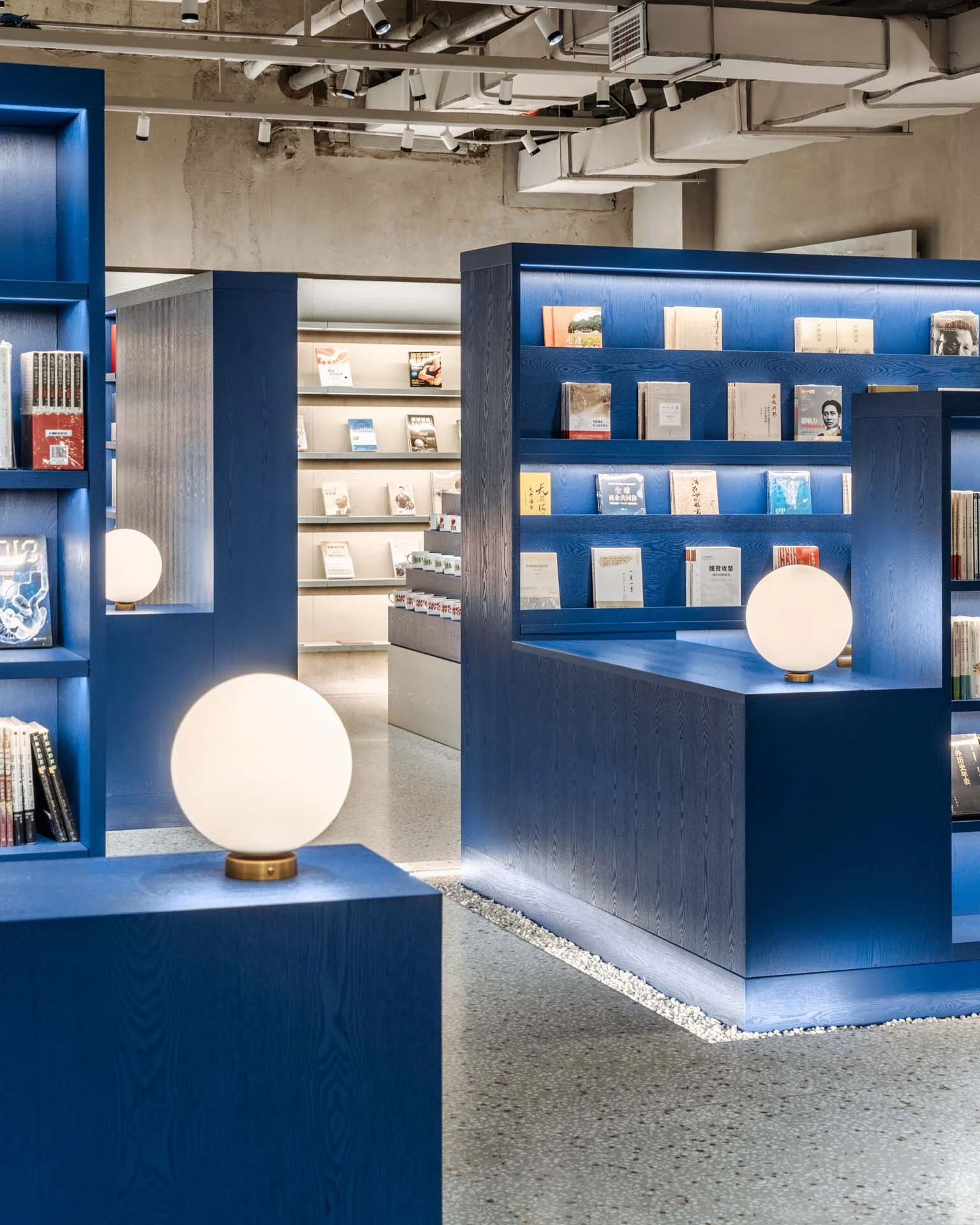 LVWA Bookstore by Studio YUDA + Studio NOR, photographed by CreatAR Images — a pair of blue-oak vitrines holds an opal-globe table lamp on a brass collar; behind them, a back room reveals a horizontal display rack carrying covers face-out.