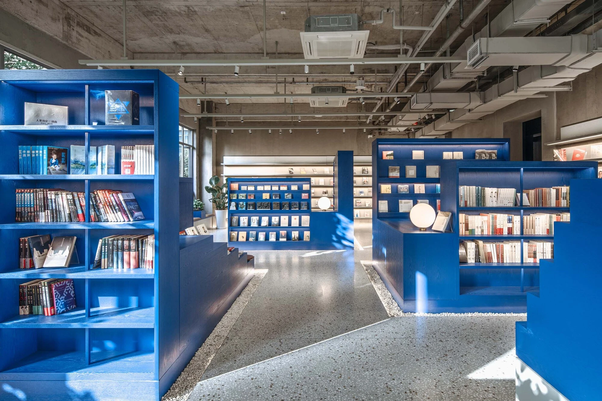 LVWA Bookstore by Studio YUDA + Studio NOR, photographed by CreatAR Images — the ground-floor sales hall: open-faced cobalt shelving units stand on a terrazzo floor under raw board-formed concrete soffits and exposed ductwork, Chinese-language paperbacks