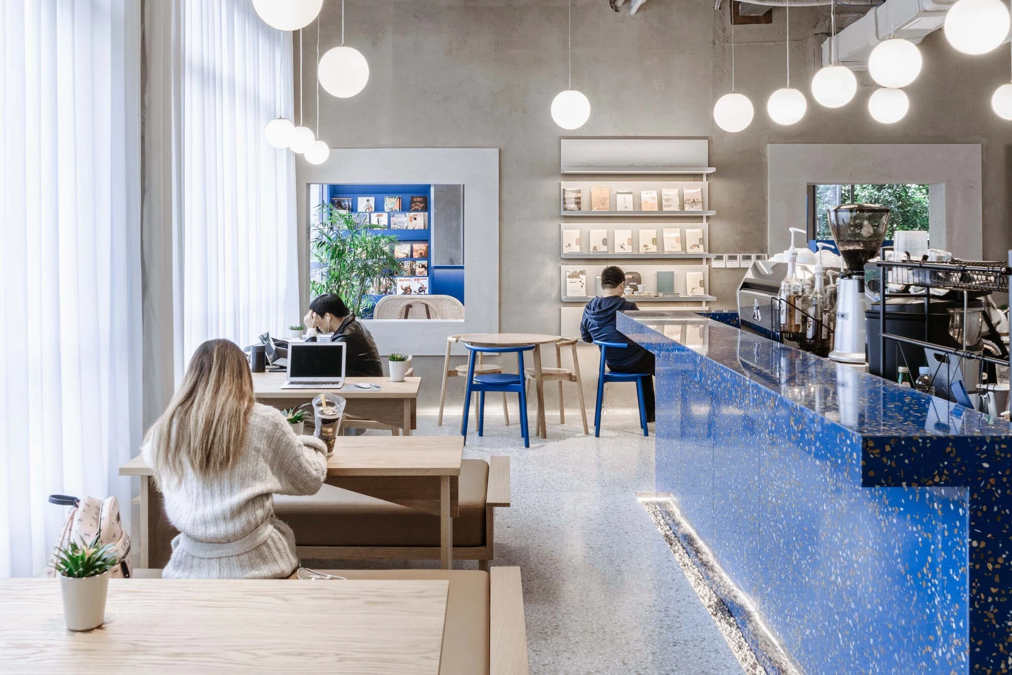 LVWA Bookstore by Studio YUDA + Studio NOR, photographed by CreatAR Images — the café bar runs in blue-and-gold terrazzo along one side; a row of suspended globe pendants picks up the same cobalt in the seated readers' high stools and tables of pale ash.
