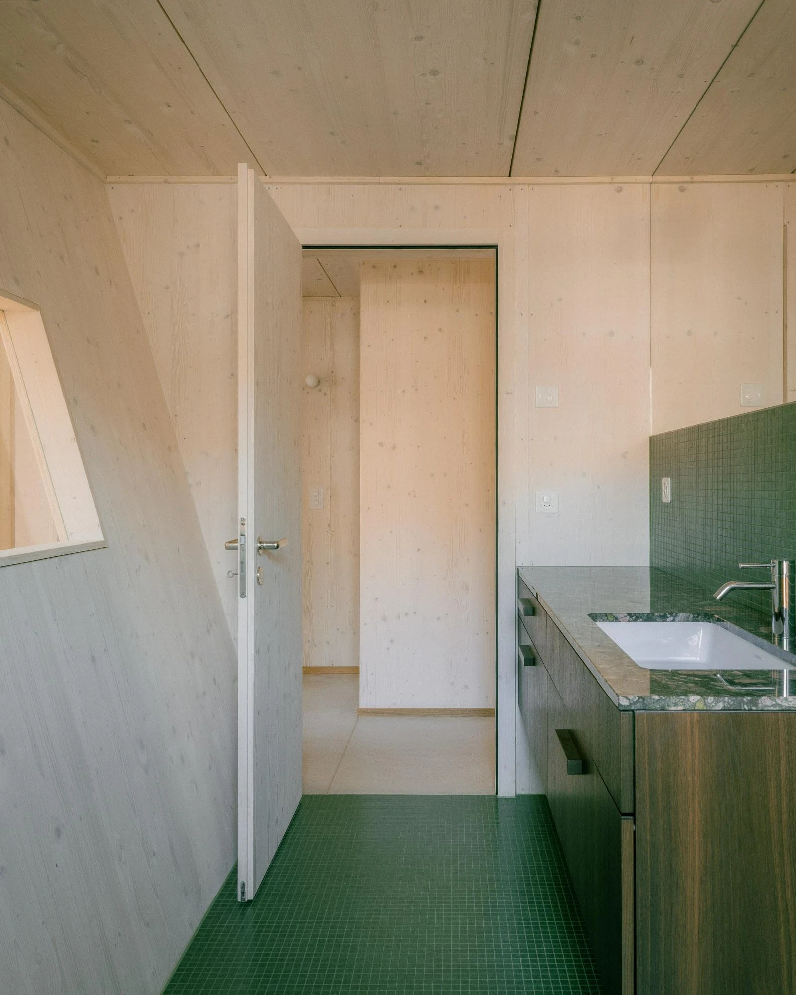 Barn Conversion in Hettlingen by Orso Dettling, photographed by Jeremiah Schwery — Bathroom in small green ceramic mosaic with a dark timber vanity and granite worktop, spruce CLT walls meeting under the gable line