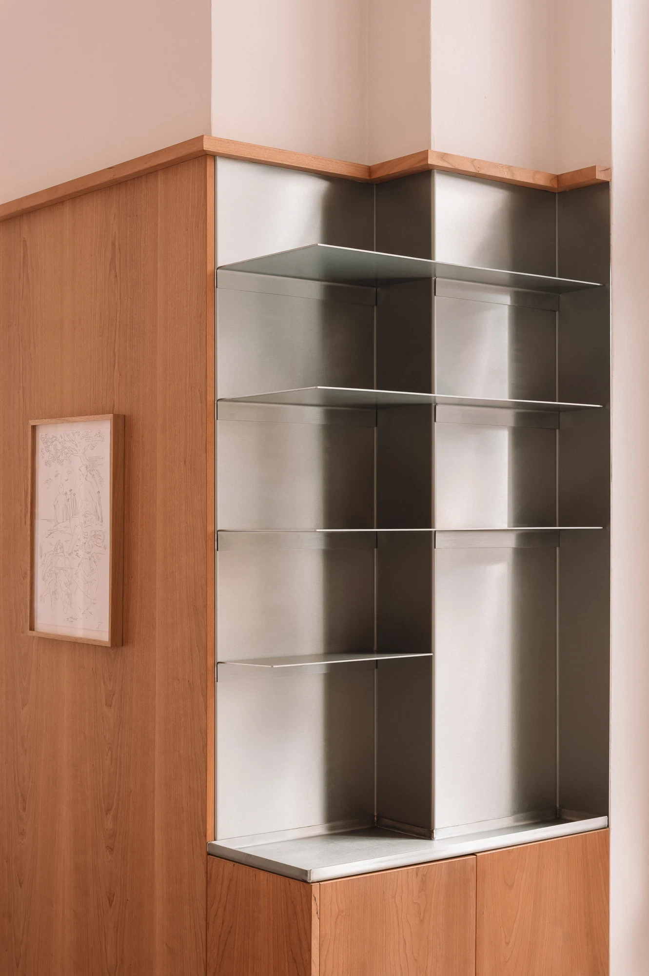 Strom Berlin by Celeste Asfour, photographed by Clemens Poloczek — A corner shelving unit in brushed stainless folds into faceted bays against cherry joinery, with a framed pencil drawing to the left.