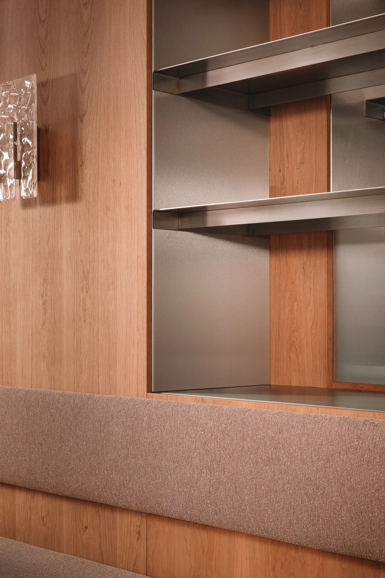 Strom Berlin by Celeste Asfour, photographed by Clemens Poloczek — A brushed stainless shelving unit with rounded brackets sits beside a Murano glass sconce on a cherry wood pillar above a mauve bouclé bench.