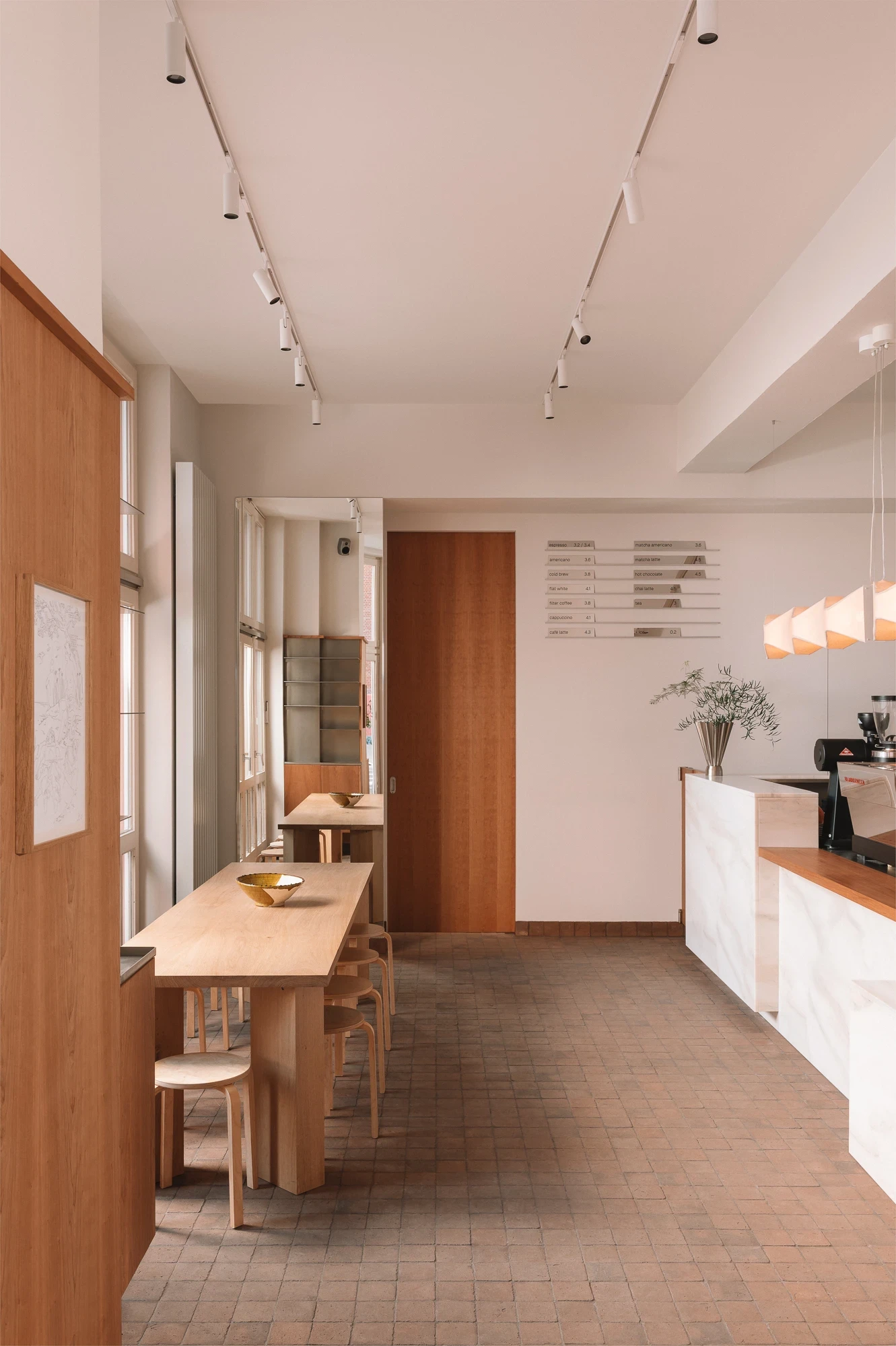 Strom Berlin by Celeste Asfour, photographed by Clemens Poloczek — Track lighting runs the length of the cafe over a long oak communal table set against terracotta paving and the marble bar in soft afternoon light.