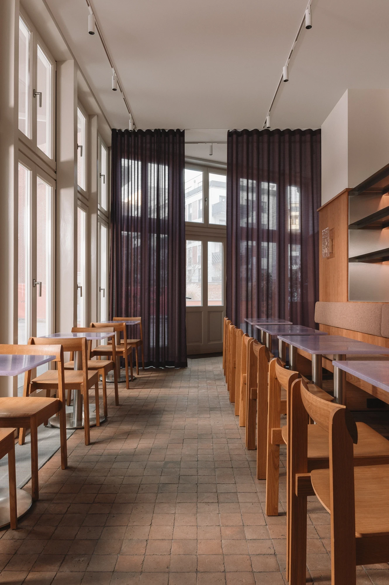 Strom Berlin by Celeste Asfour, photographed by Clemens Poloczek — Sheer aubergine curtains run floor to ceiling across the courtyard windows above a row of light oak chairs and lavender resin tables.
