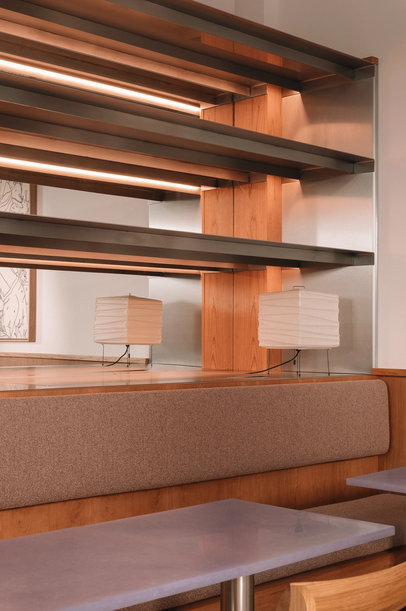 Strom Berlin by Celeste Asfour, photographed by Clemens Poloczek — Paper lanterns sit on either side of a low cherry partition above the banquette, illuminated by warm strips tucked beneath each steel shelf.