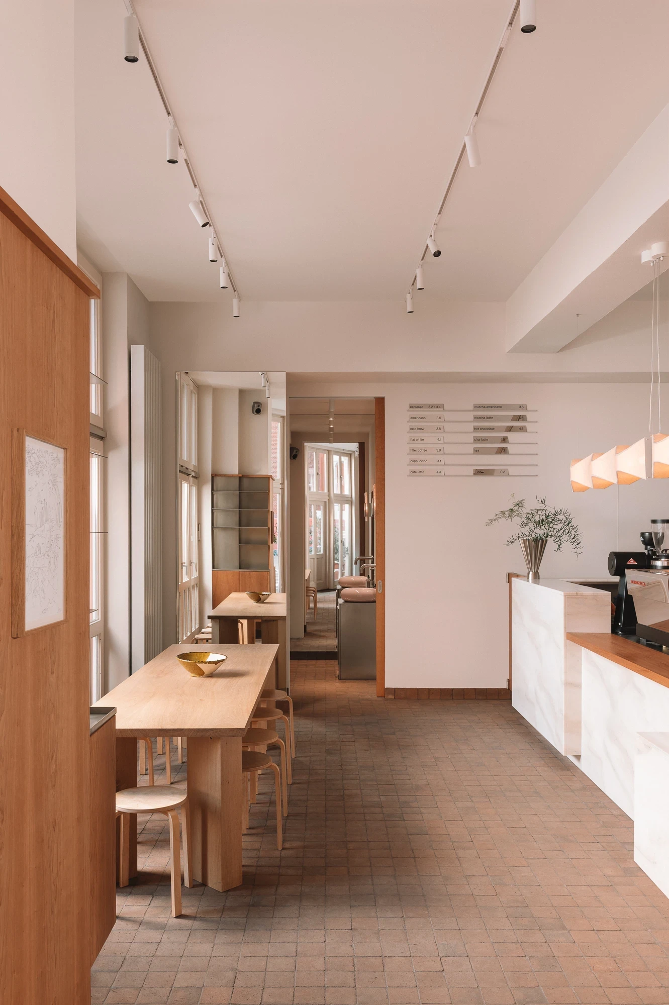Strom Berlin by Celeste Asfour, photographed by Clemens Poloczek — A view down the cafe shows the marble bar, oak tables, and printed menu strips on the back wall with light coming through the courtyard windows.