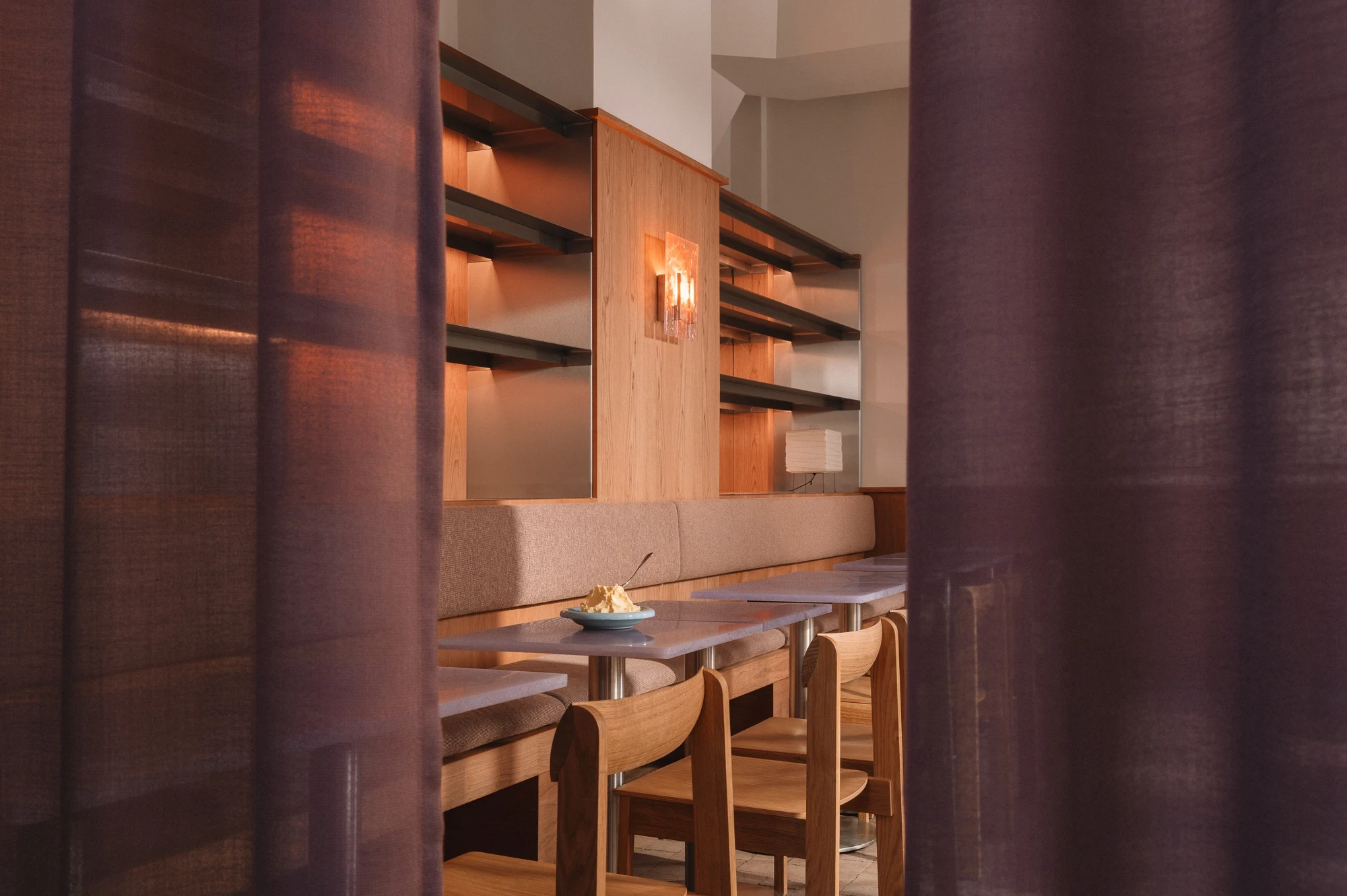 Strom Berlin by Celeste Asfour, photographed by Clemens Poloczek — An interior view past a curtain edge shows the long banquette wall, a paper lantern beside a glass sconce, and a small bowl of ice cream on the table.